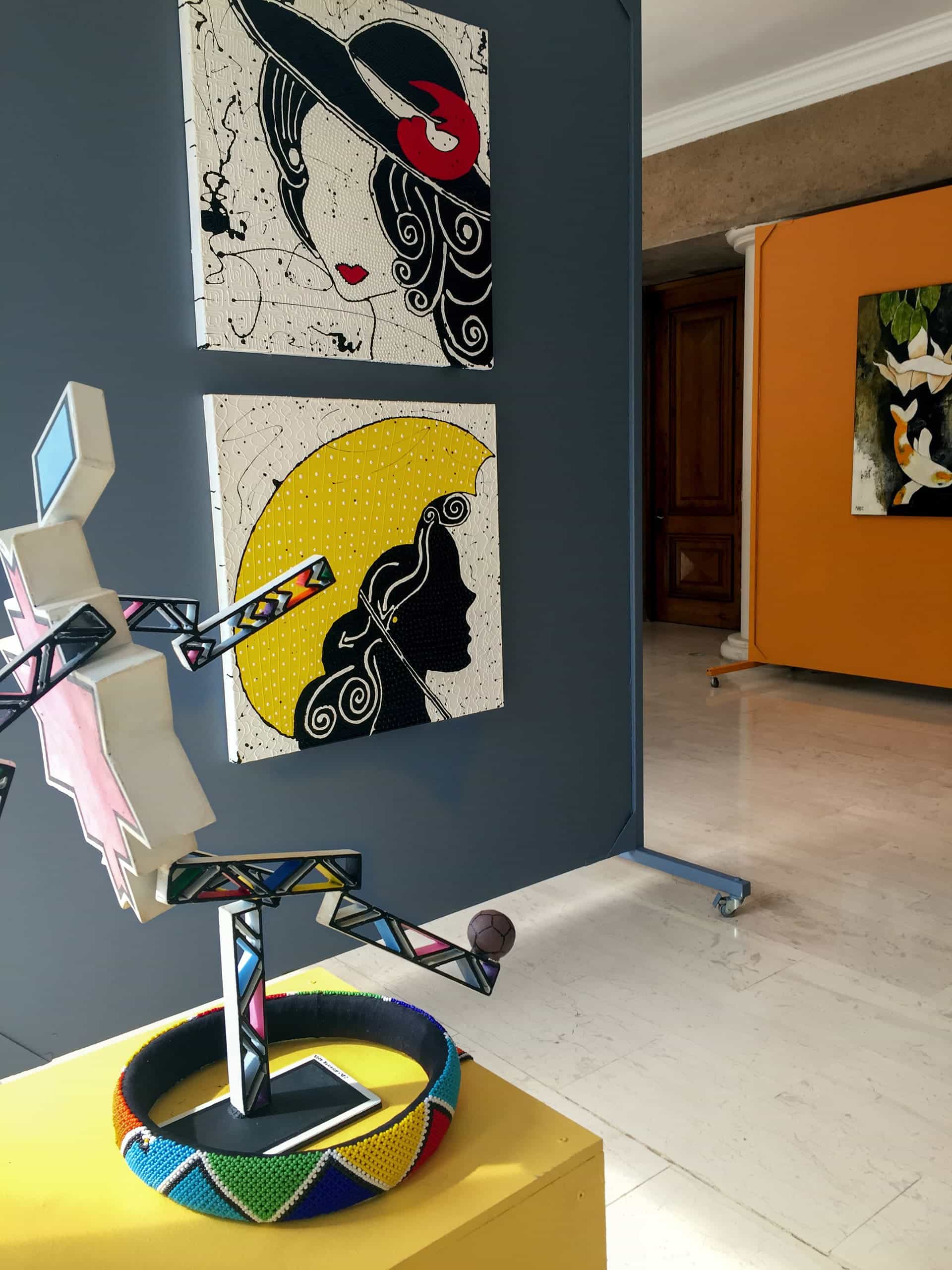 A modern art gallery features colourful geometric sculptures, bold graphic paintings of women in hats, and abstract art on orange and grey walls. A beaded sculpture stands on a yellow plinth in the foreground, with paintings displayed on panels behind it.