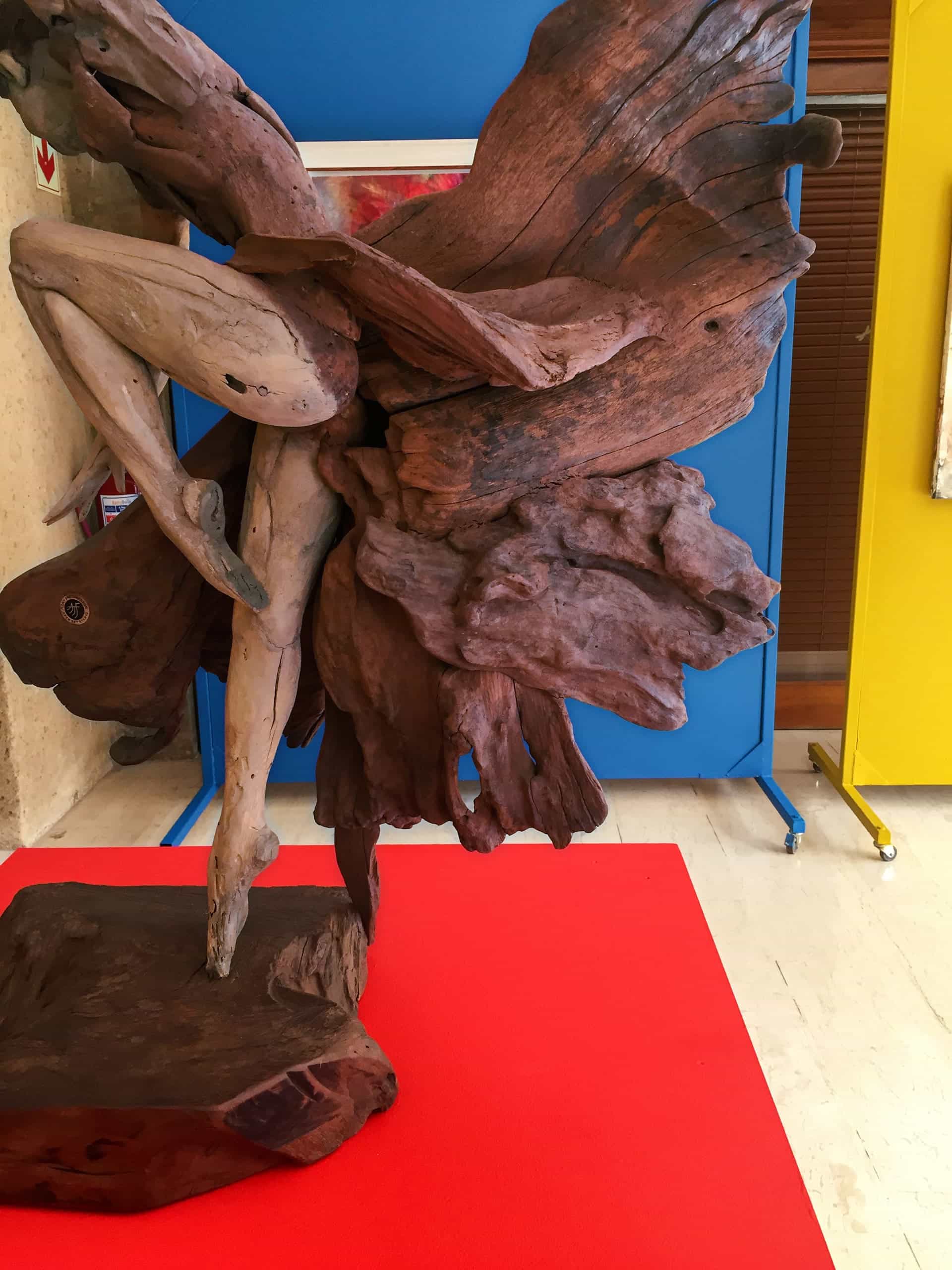 A wooden sculpture made from large, twisted driftwood pieces forms an abstract figure resembling a dancer or fairy with outstretched limbs and wing-like shapes. The sculpture stands on a red mat against blue and yellow display panels indoors.