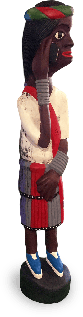 A painted wooden figurine of a woman with dark skin, wearing a colourful skirt, red scarf, blue shoes, and metal bangles. She holds a black mobile phone to her ear, and her hair is covered by a green and red headscarf.