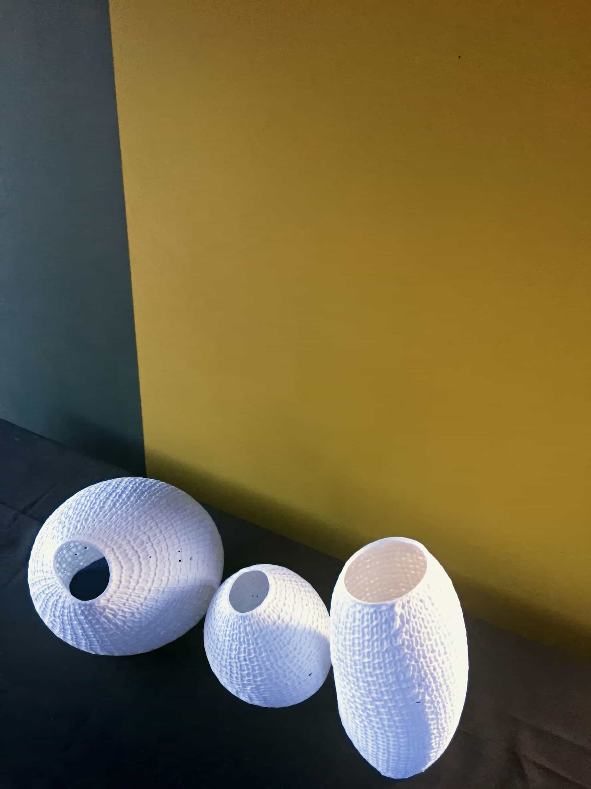 Three white, textured, oval vases of varying heights are arranged on a dark surface. The background is divided vertically, with the left side a muted green and the right side a mustard yellow. Bright light highlights the vases' patterns.