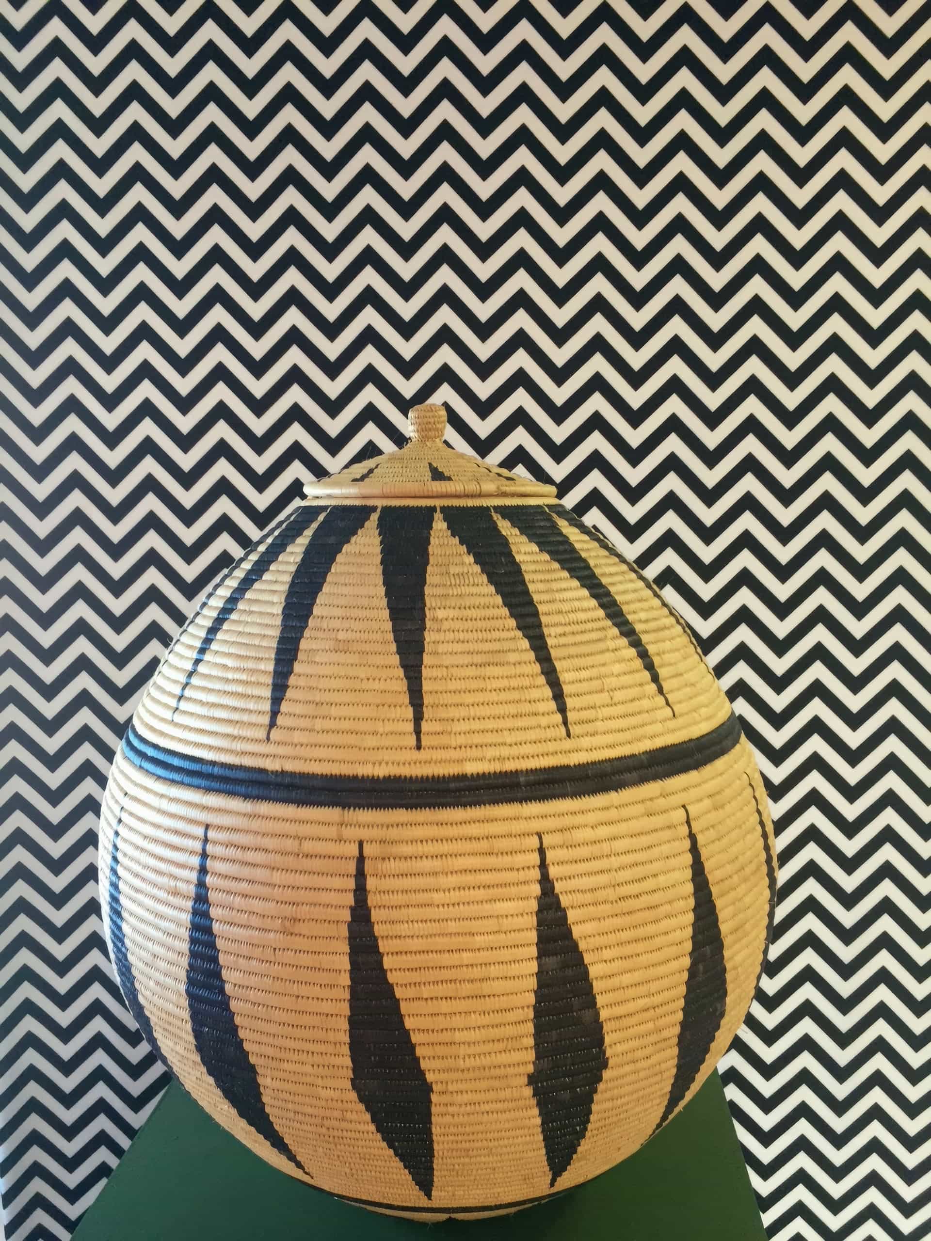 A woven, round lidded basket with a geometric black and natural pattern sits on a green surface. The background is a bold black and white zigzag chevron wallpaper, creating a striking contrast with the basket’s design.