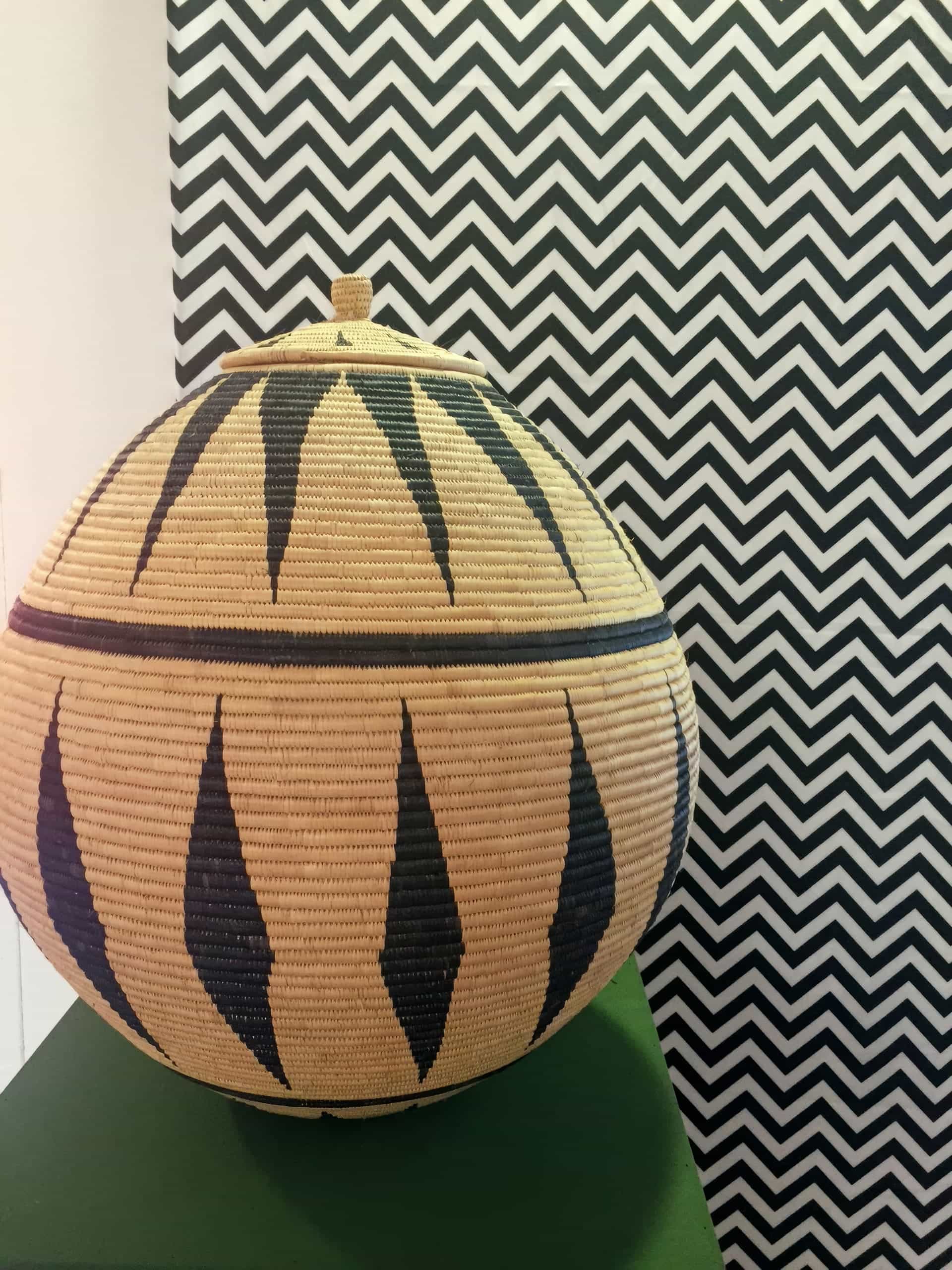 A large, round woven basket with a lid and geometric black patterns sits on a green surface. The background features a bold black and white chevron zigzag pattern. The basket’s lid has a small handle on top.