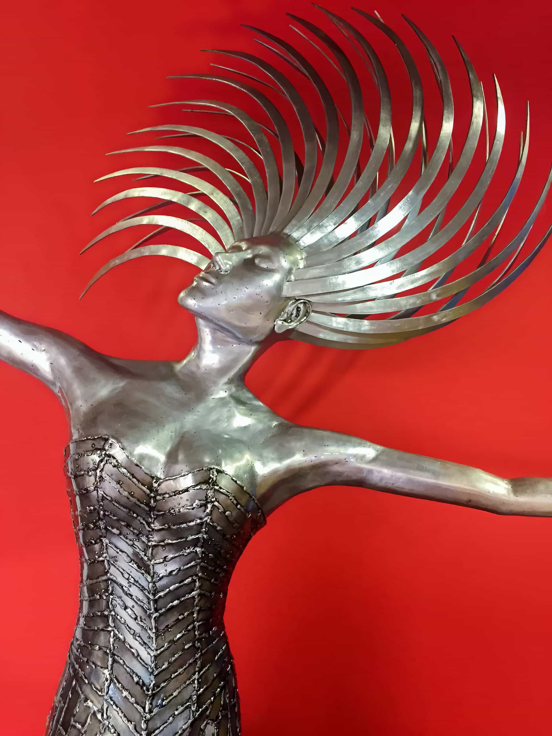 A metallic sculpture of a female figure with arms outstretched, wearing a detailed bodice and a dramatic headdress made of curved, radiating metal spikes. The background is solid bright red, creating a striking contrast with the silver tones of the sculpture.