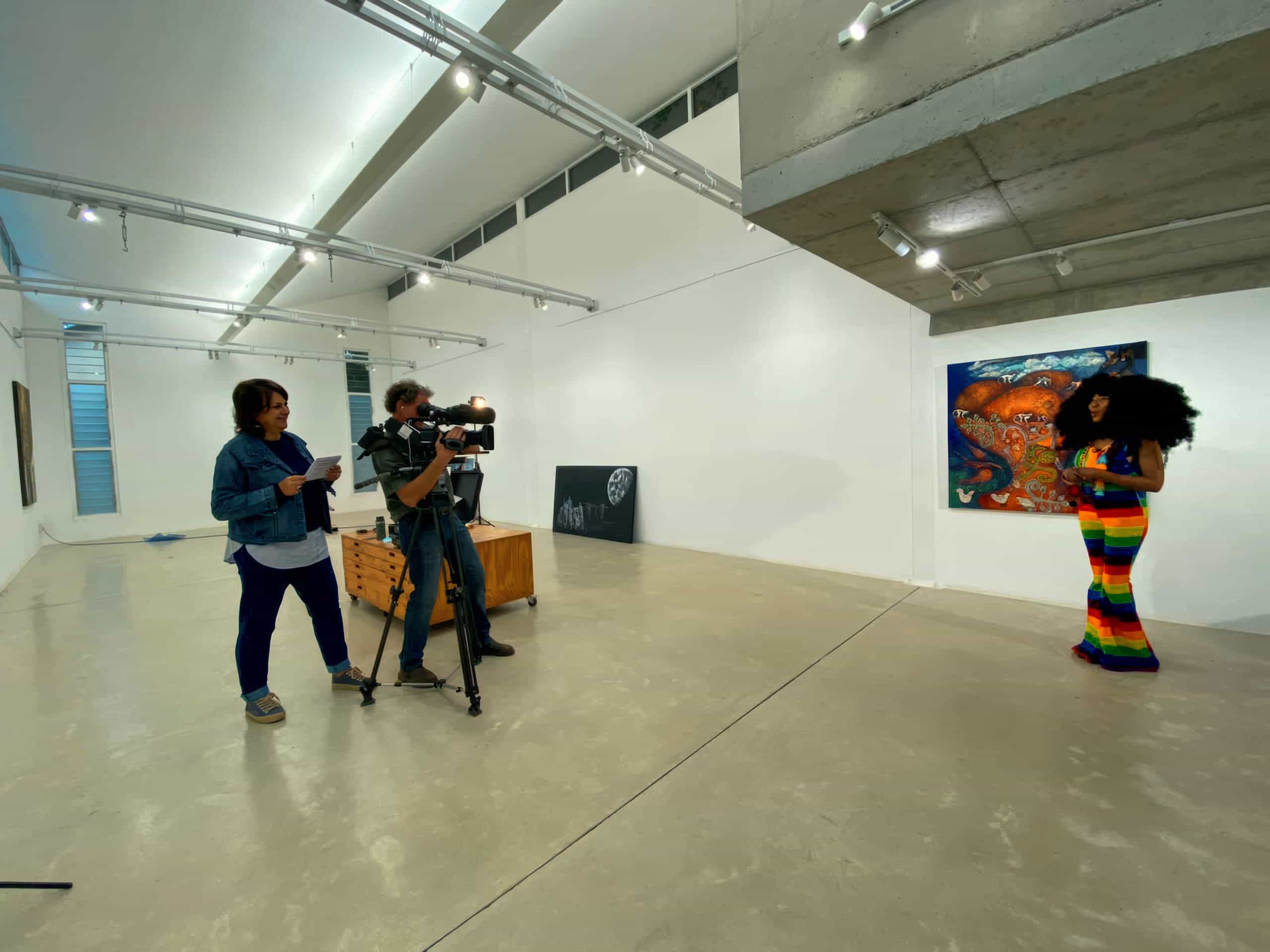 In a modern art gallery, a camera crew films a woman in a colourful rainbow outfit presenting in front of a vivid painting; artworks hang sparsely on white walls, and bright lights illuminate the open, polished concrete space.