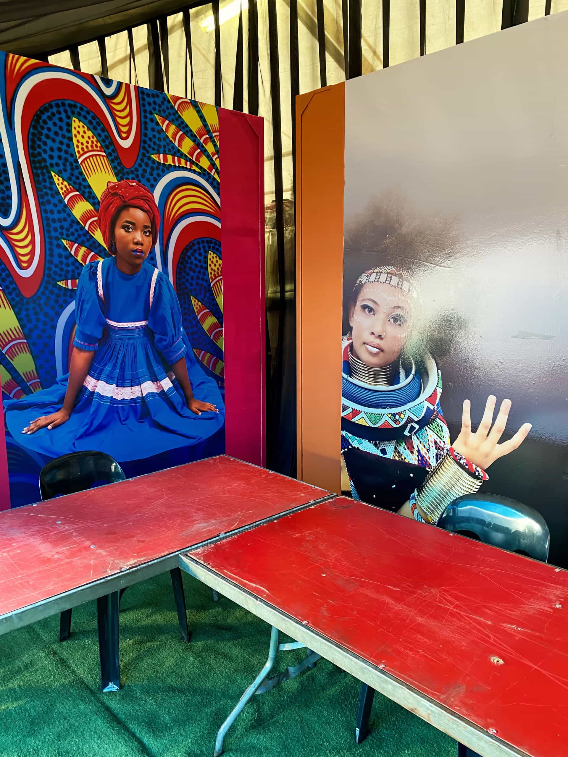 Two colourful portraits of Black women are displayed behind red tables and black chairs. One woman wears a blue dress and patterned headwrap, seated before a vibrant abstract background. The other is in futuristic attire, raising her hand in a gesture.
