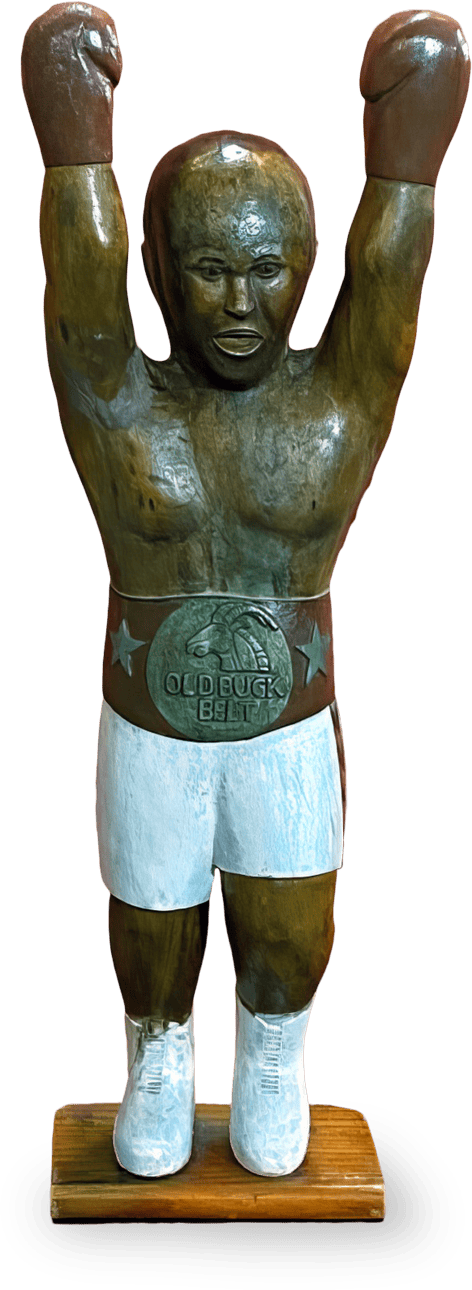 A wooden sculpture of a boxer with arms raised triumphantly. The figure wears white boxing shorts, white shoes, and a large silver belt reading “OLD DOG BELT.” The sculpture is mounted on a rectangular wooden base.