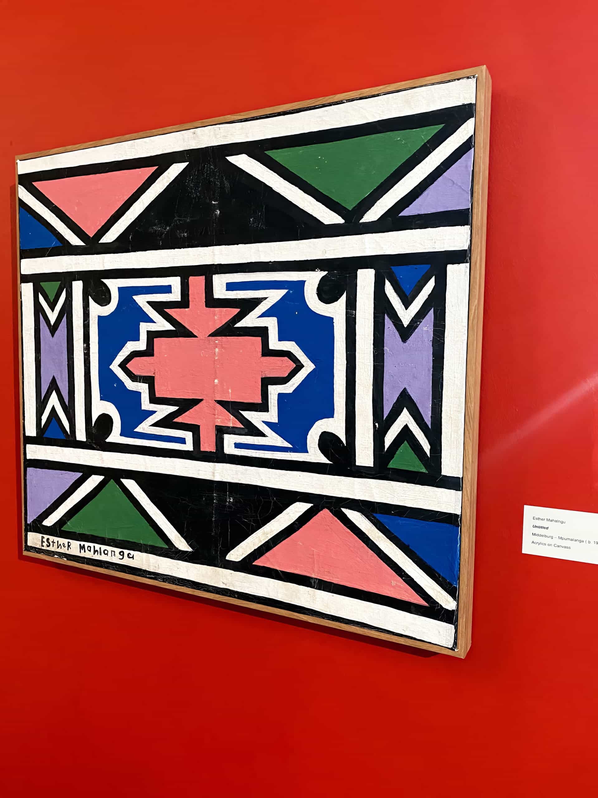 A geometric painting in bold colours—blue, pink, green, black, and white—featuring symmetrical patterns and shapes, hangs on a vibrant red wall. The artist’s signature, Esther Mahlangu, is visible in the lower left corner. A small label is mounted nearby.