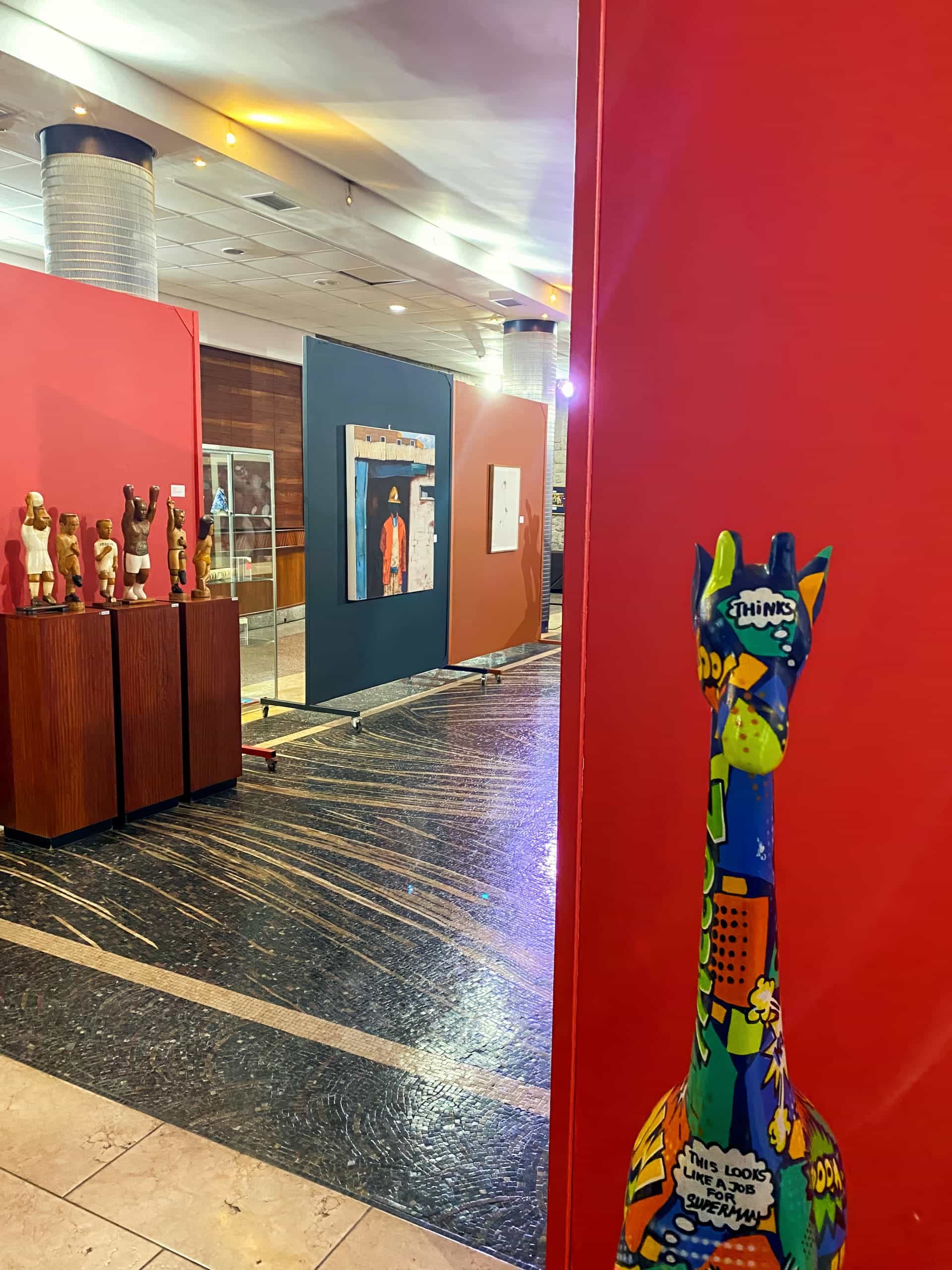 A colourful, comic-themed giraffe sculpture stands near a red wall at an art gallery. In the background, sculptures and framed artworks are displayed on stands and panels under warm lighting, with a glossy floor and modern decor visible.
