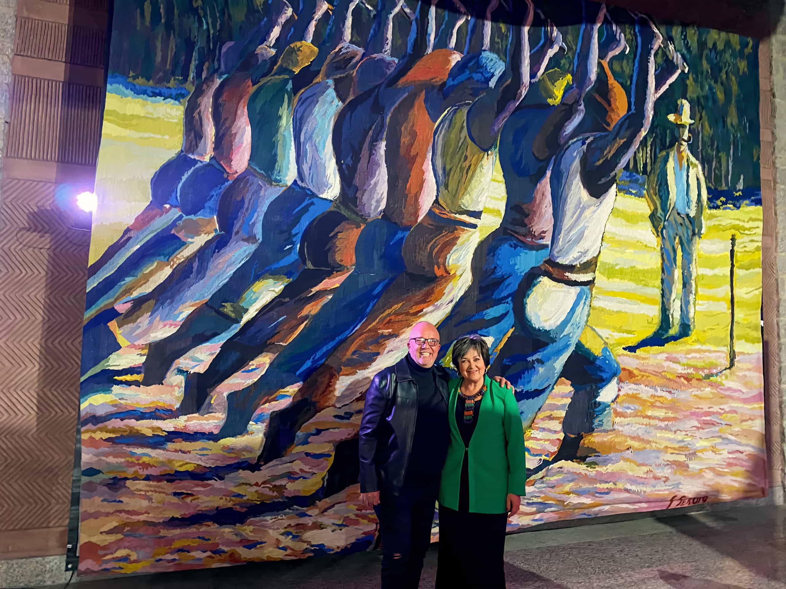 Two people stand smiling in front of a vibrant mural. The mural depicts a group of workers, arms raised, painted in bold colours. A single figure stands off to the right. The people in front are well-dressed and posed together under bright lighting.