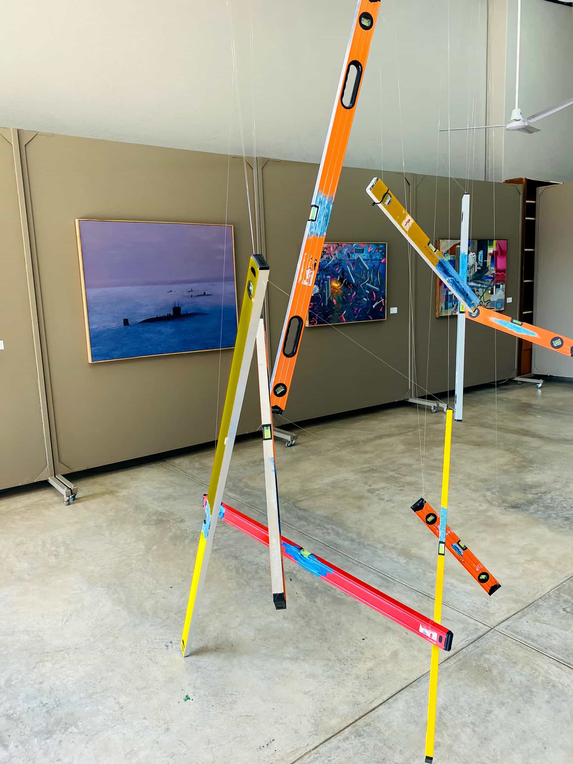A sculpture made of colourful spirit levels suspended by thin wires stands on a concrete floor in an art gallery. Behind it, paintings hang on beige walls, including a seascape and abstract artworks. The space is bright and modern, with high ceilings.