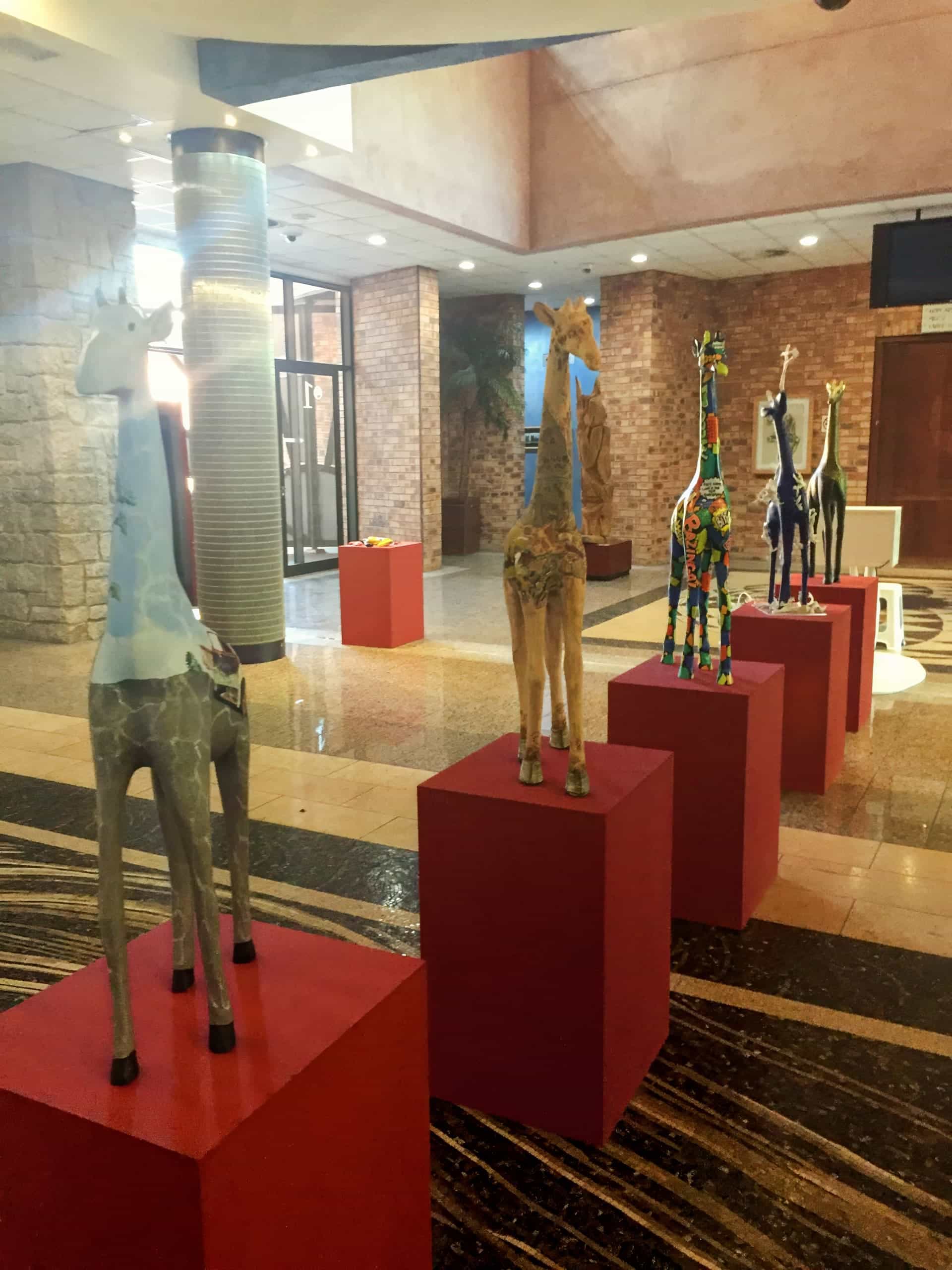 Five colourful giraffe sculptures stand on red plinths in a spacious, well-lit lobby with marble floors, brick walls, and large windows. Each giraffe features a unique pattern and design, contributing to a vibrant, artistic atmosphere.