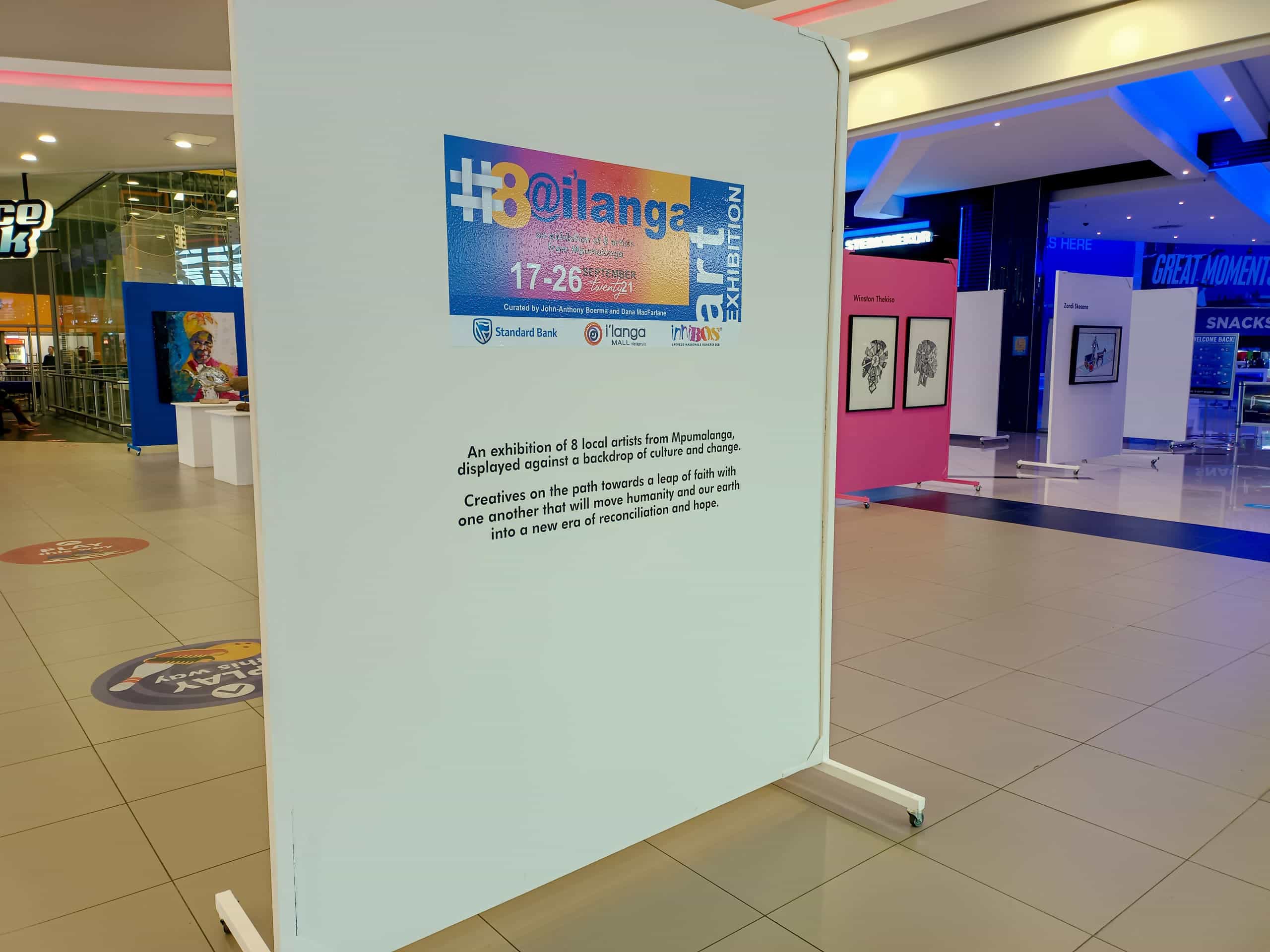 A white exhibition panel in a shopping centre describes the “#30@ilanga” art exhibition, with logos at the top. The space is open, with framed artworks on easels in the background and shopfronts visible, including a sign for GREAT MOVIES.
