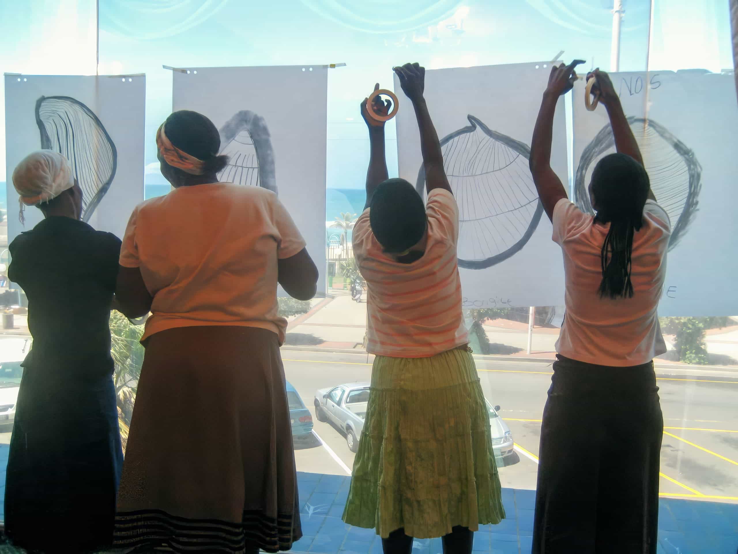 Four people stand indoors facing large windows, hanging up drawings of seashells on sheets of white paper. Sunlight streams in, casting their silhouettes. Outside, cars, palm trees, and the sea are visible through the glass.