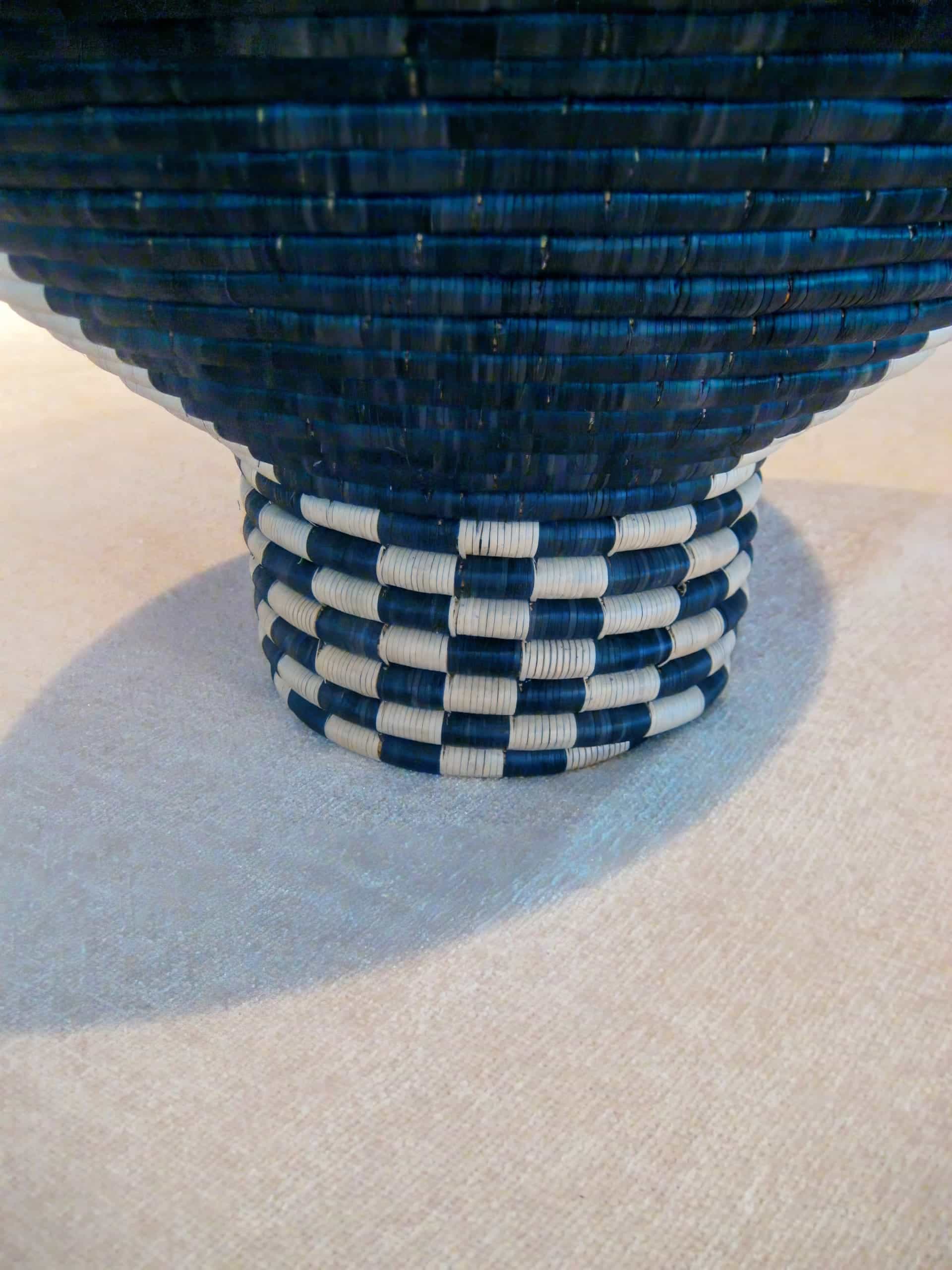 A close-up of the base of a woven basket with dark blue and white alternating bands in a chequered pattern. The surface underneath is a light beige fabric, and the shadow of the basket is visible on the left side.