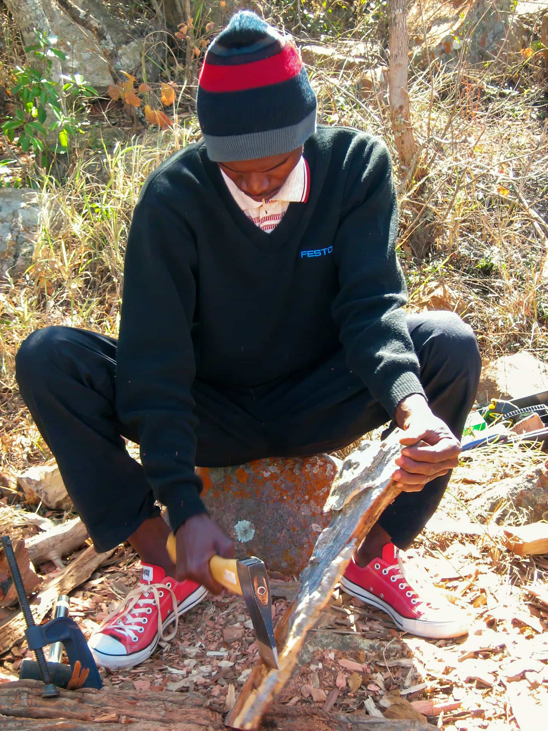 A person wearing a black jumper, red and black hat, red trainers, and a white mask saws a piece of wood while sitting outdoors on a rock surrounded by dry grass and leaves. The sunlight highlights the earthy, natural setting.