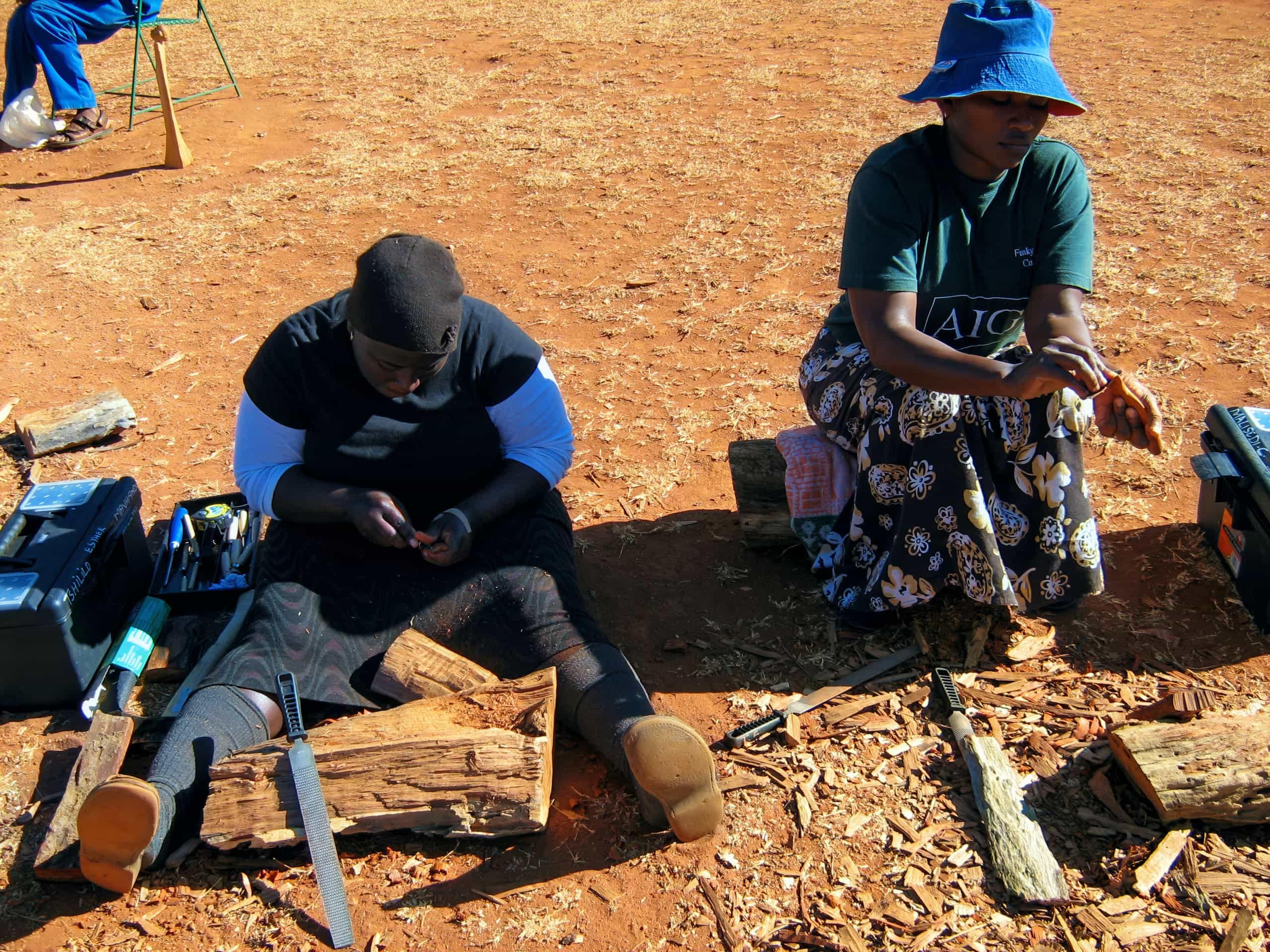 Two women sit on the ground outdoors on reddish earth, working with tools and wood. One woman is filing or carving a piece of wood, while the other holds a tool. Logs, toolboxes, and wood shavings are scattered around them. Both wear hats and long skirts.