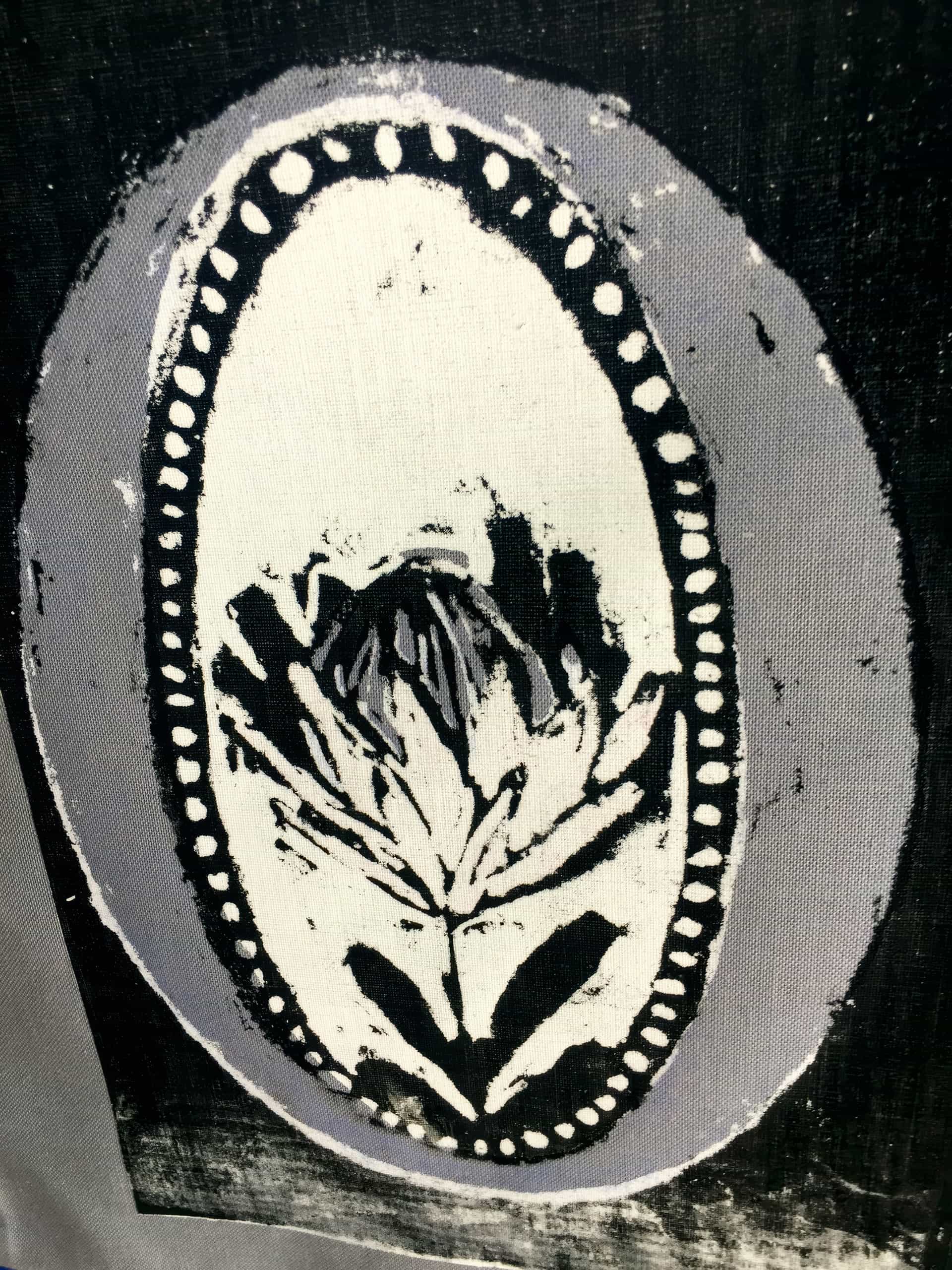 A black and white fabric print features an oval frame with a dotted border. Inside the oval, there is a stylised, abstract flower with bold, dark brushstrokes on a light background. The outer edges are shaded in grey, giving a textured appearance.