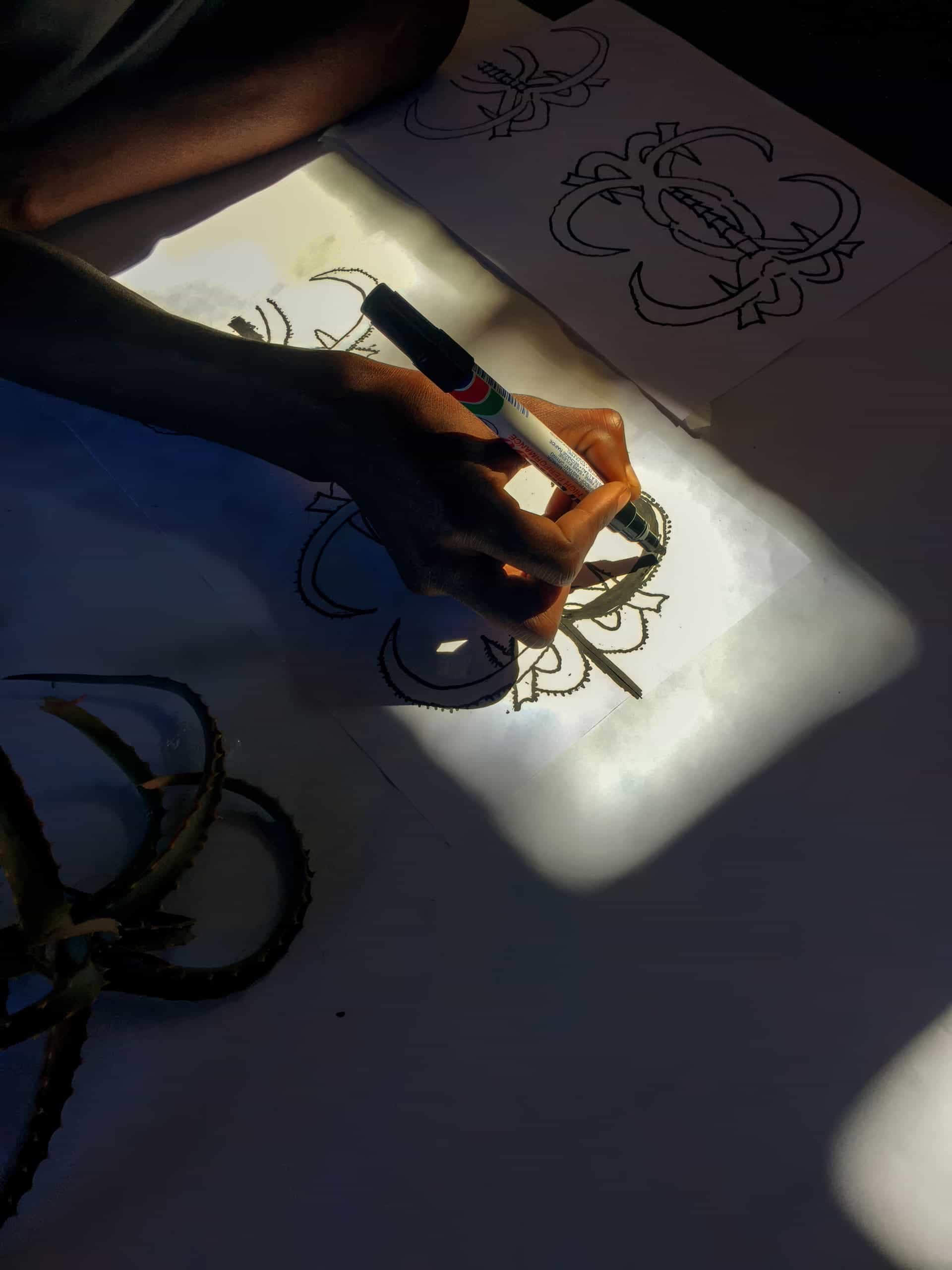 A hand holding a black marker traces over a detailed floral line drawing on white paper, illuminated by a beam of sunlight. Other similar sketches and part of a plant with long, curved leaves are visible nearby in the shadow.