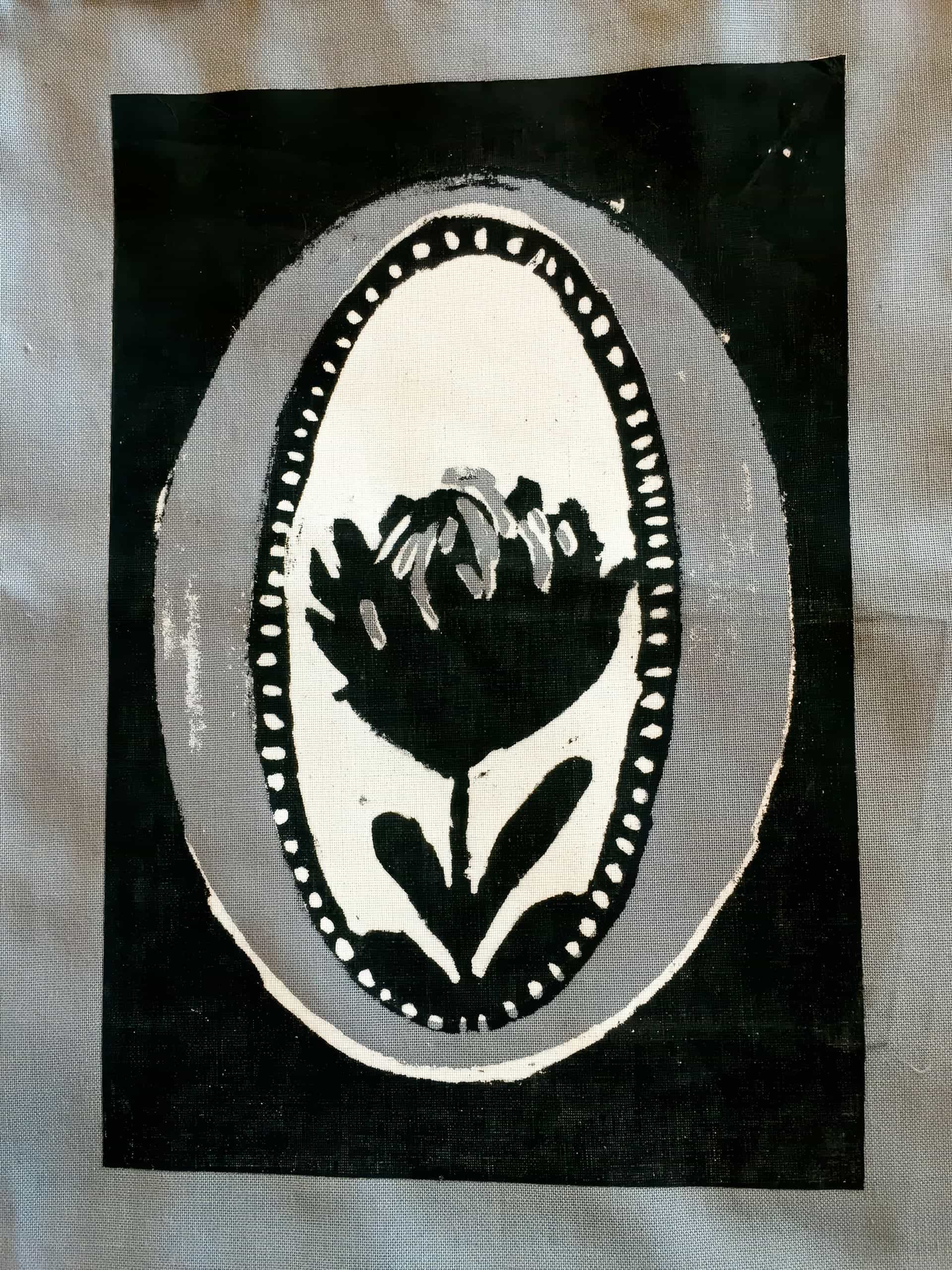 A black and white print depicts a stylised flower with two leaves inside an oval border. The border is decorated with small dots. The flower and borders are surrounded by a solid black rectangular background, set against grey fabric.
