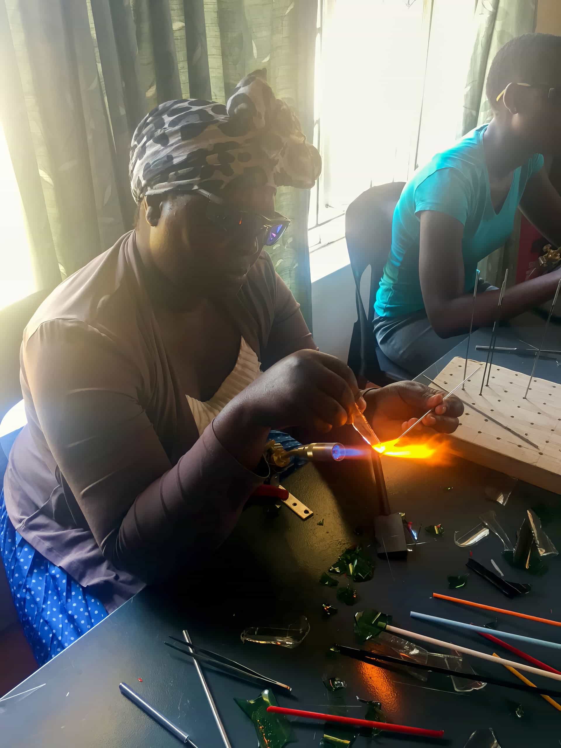 A person wearing glasses and a patterned headscarf shapes glass using a blowtorch, creating an orange flame. Glass pieces, tools, and coloured glass rods are scattered on the table. Another person works in the background. Sunlight filters through curtains behind them.