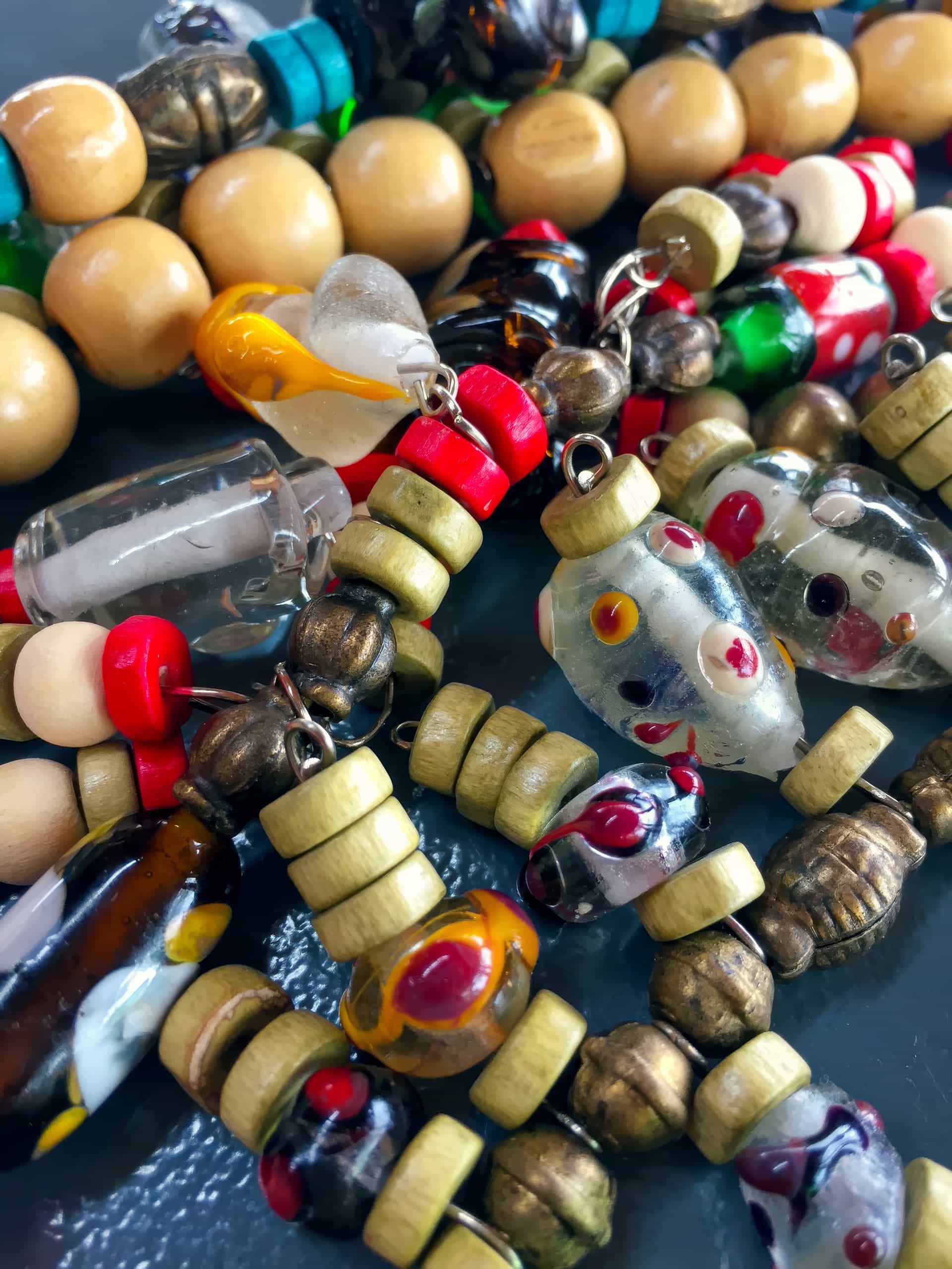 A close-up of colourful beaded necklaces featuring round wooden, metallic, and glass beads, including clear glass beads with red and yellow patterns, green and red beads, and some with heart and capsule shapes, all arranged in a tangled pile.