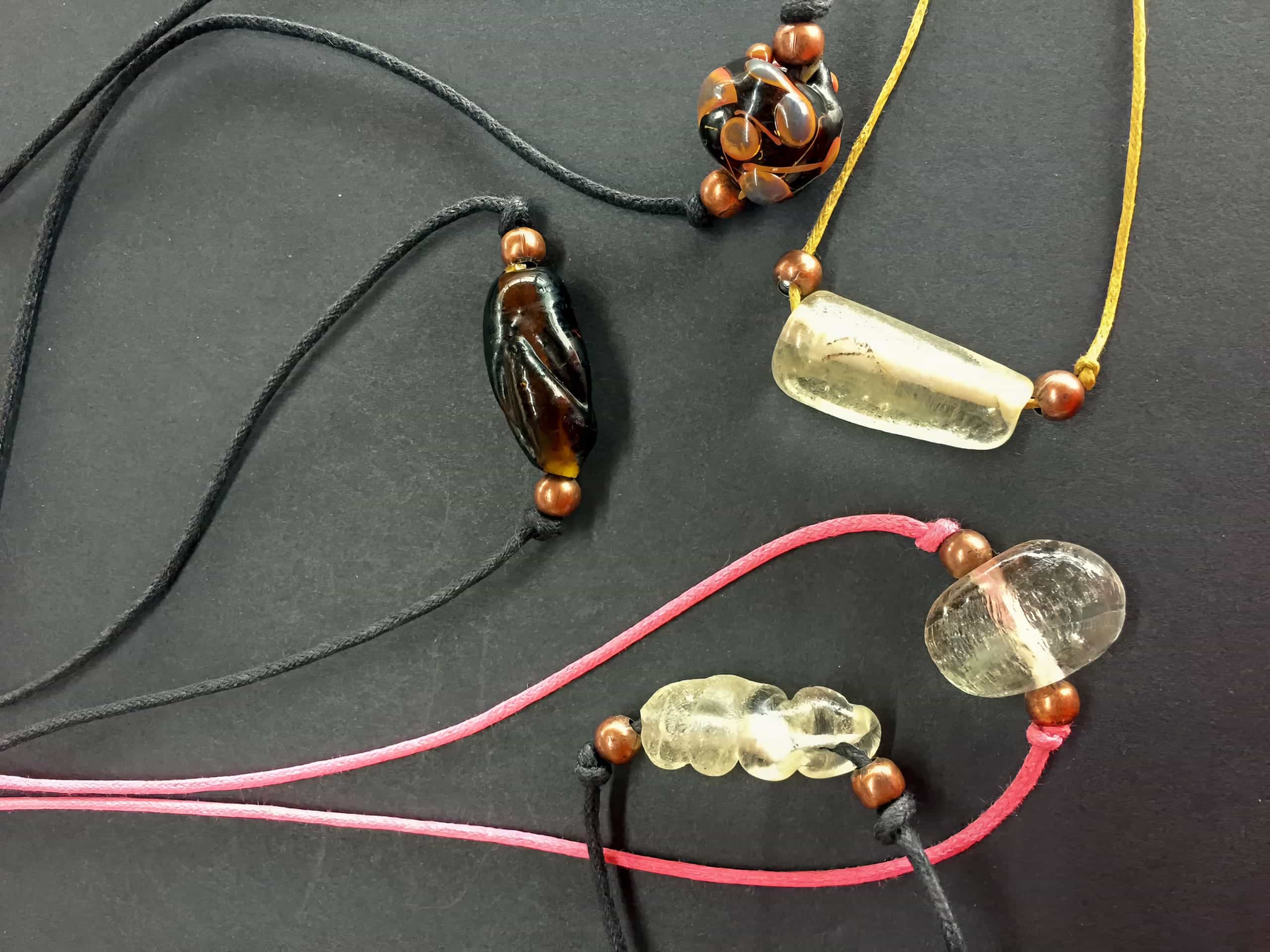 Four handmade necklaces with black, yellow, and pink cords, each featuring a unique polished stone or glass pendant and wooden bead accents, are arranged on a black surface. The pendants vary in colour, including brown, clear, and amber tones.