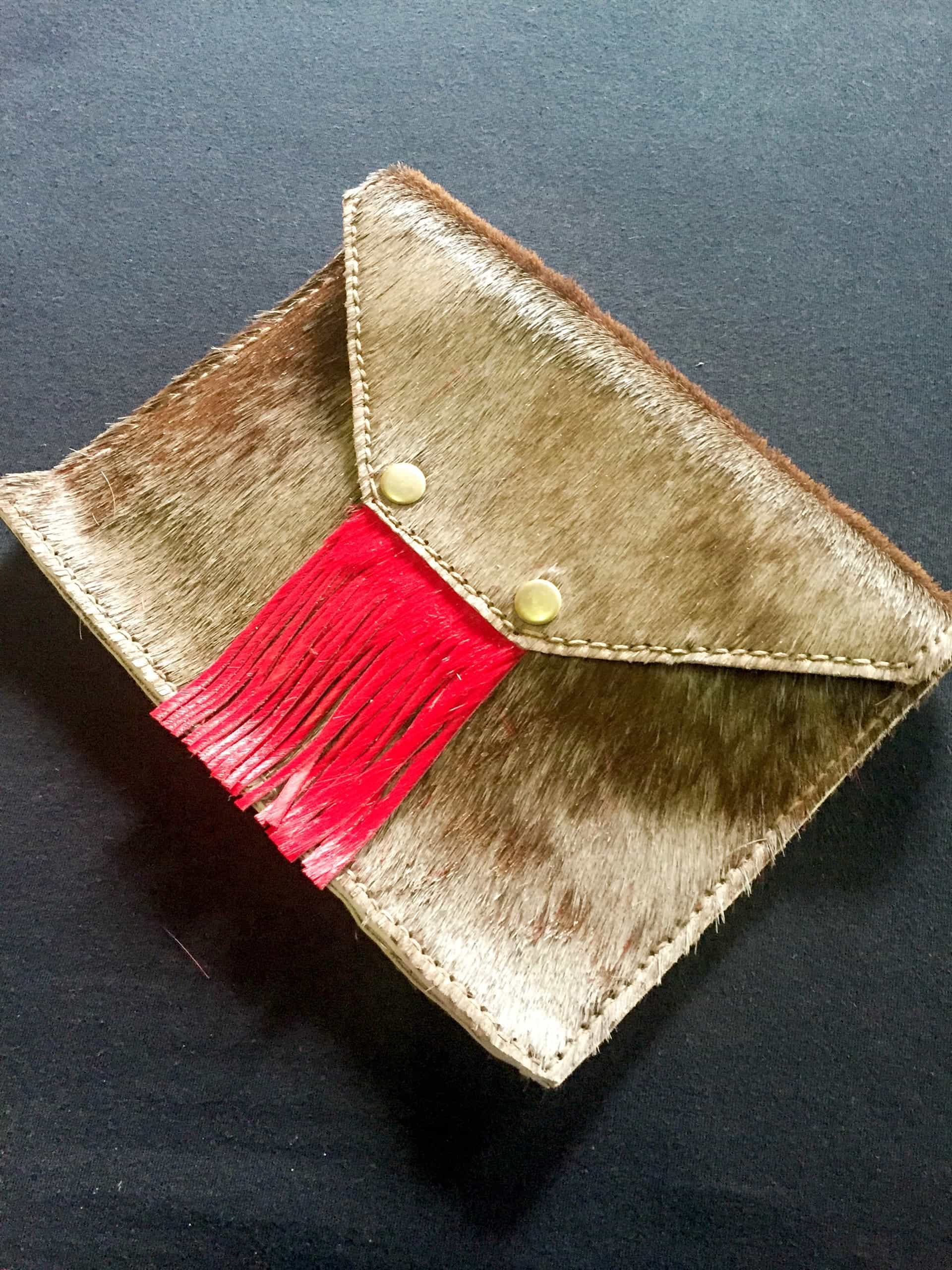 A rectangular brown and beige fur clutch bag with a pointed flap, gold stitching, two gold press studs, and red leather fringe hanging from under the flap. The bag is resting on a dark surface.