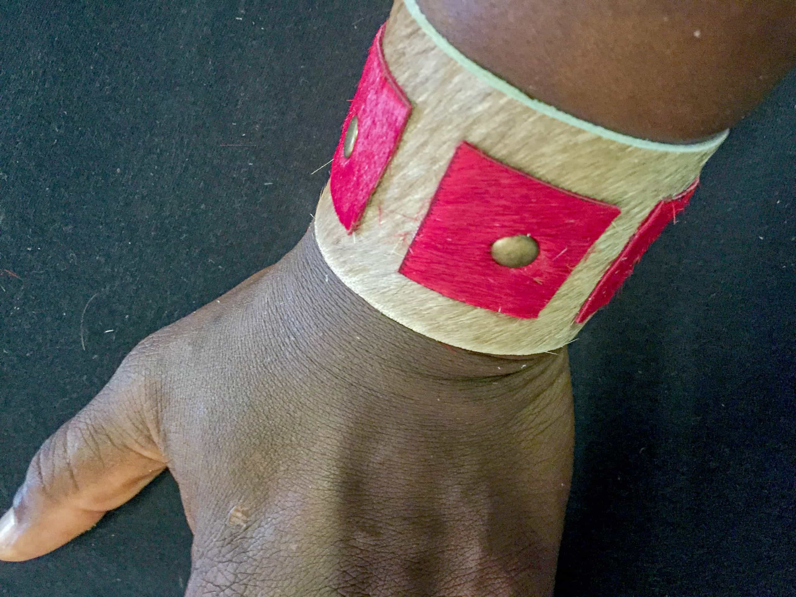 A close-up of a wrist wearing a wide, tan-coloured bracelet with red square decorations and small round metal studs, set against a black background. The bracelet features a textured appearance and contrasts with the wearer’s dark skin tone.