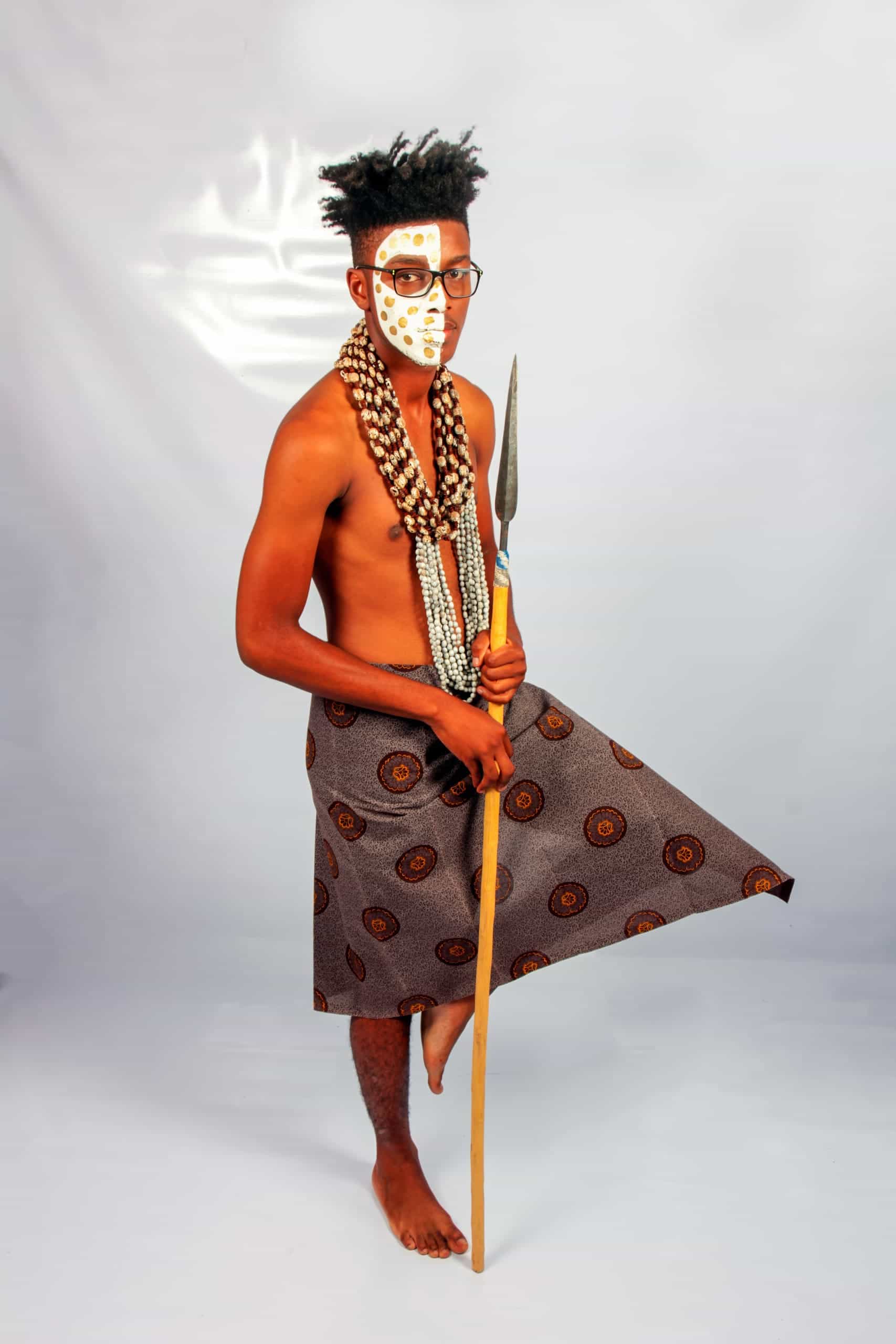 A barefoot man stands on one leg against a plain backdrop, wearing a patterned wrap skirt, layered beaded necklaces, and traditional white face paint. He holds a spear in one hand and gazes forward, embodying traditional African attire and cultural heritage.