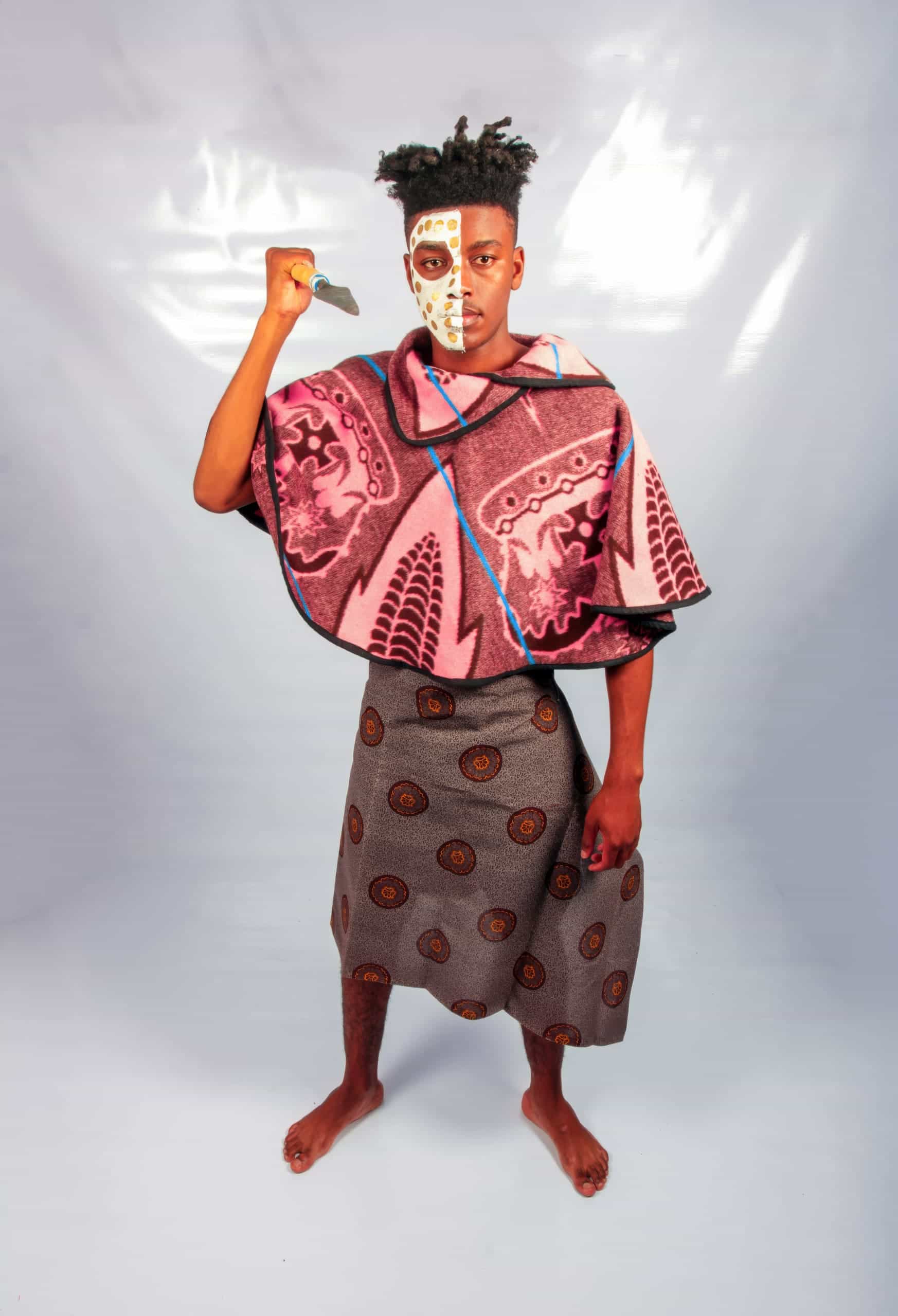 A barefoot person stands on a light floor, wearing patterned traditional clothing: a pink-and-purple poncho and a grey skirt with circular designs. They hold a knife upright, and half their face is painted white with distinct markings. The background is plain and light-coloured.