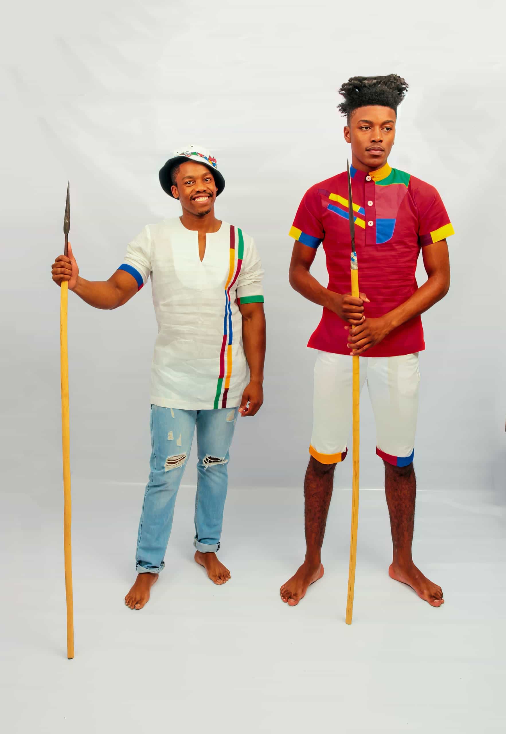 Two barefoot men stand on a white background with spears. The left man wears ripped jeans, a white shirt with colourful stripes, and a hat, smiling. The right man wears a maroon shirt and white shorts with colourful trims, looking serious.