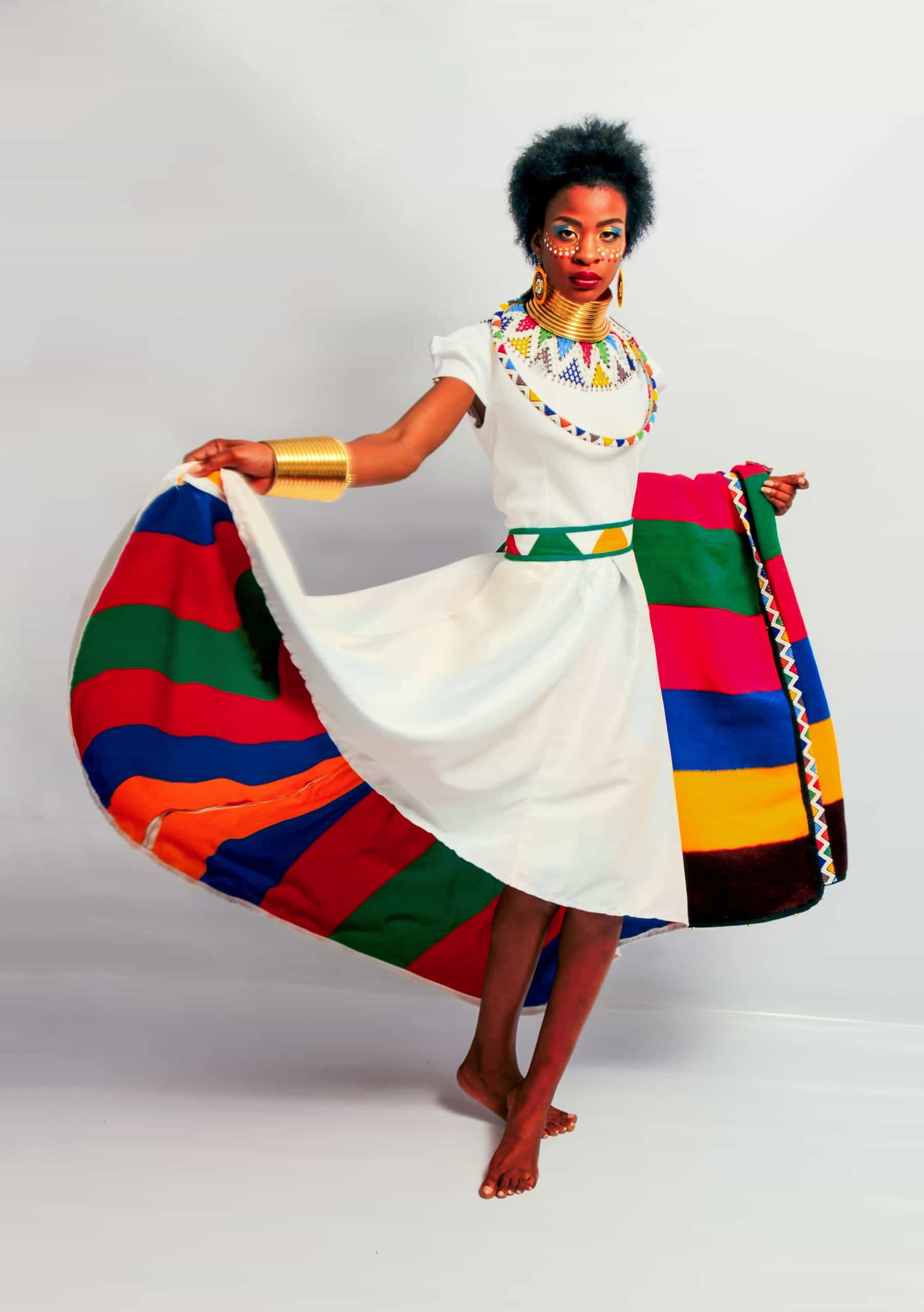 A barefoot woman poses in a white dress with colourful stripes on the skirt. She wears bold beaded jewellery, gold bangles, and traditional face paint. Her hair is natural, and she holds out her skirt to show off the vibrant design against a plain background.