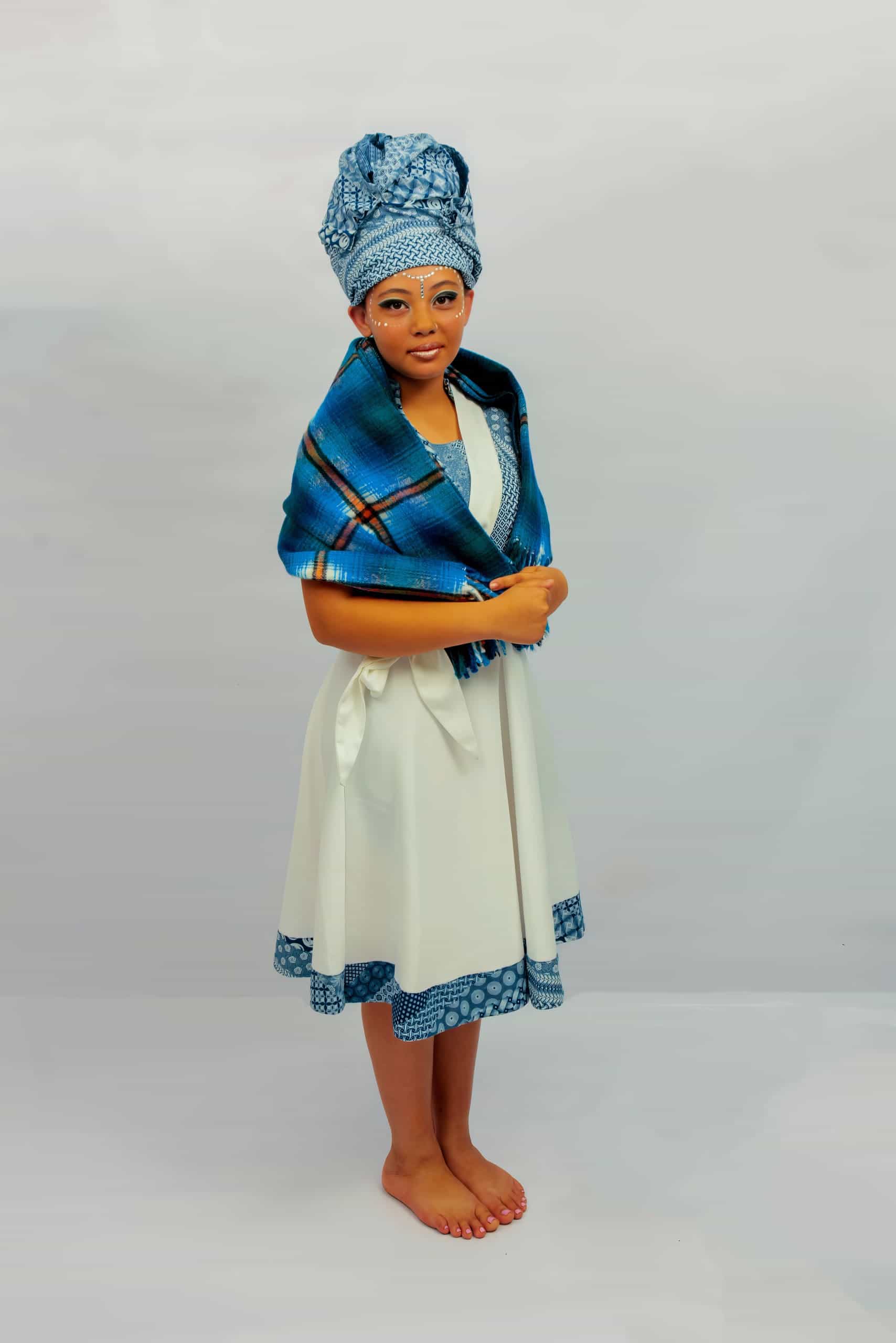 A young woman stands barefoot on a plain background wearing a white dress with blue patterned trim, a matching blue patterned headwrap, and a blue plaid shawl draped over her shoulders. She faces forwards with her hands lightly holding the shawl.
