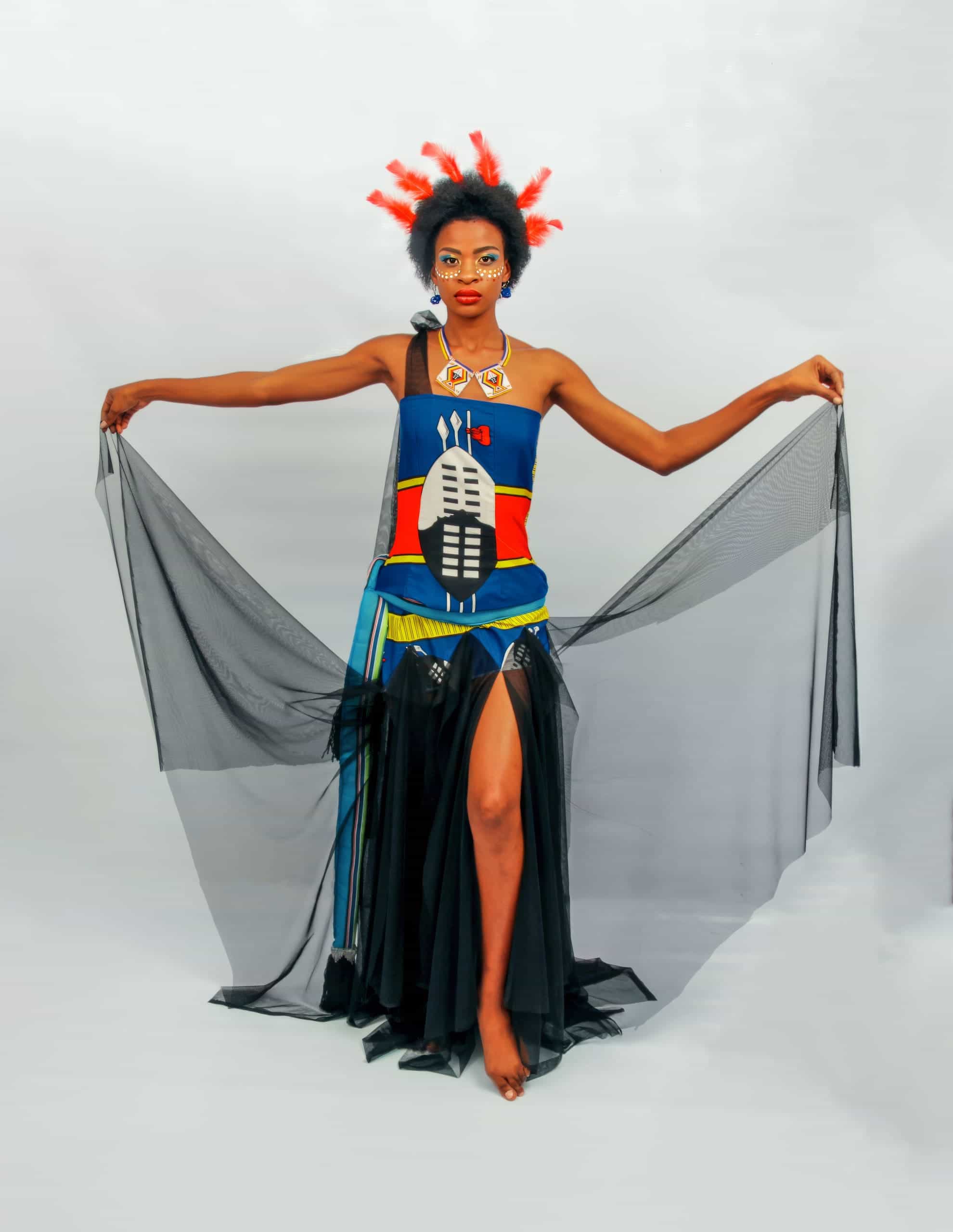 A woman stands with arms outstretched, holding sheer black fabric. She wears a colourful dress with bold patterns and a tall headdress decorated with red accents. Her make-up is bright and artistic, and she poses barefoot against a plain white background.