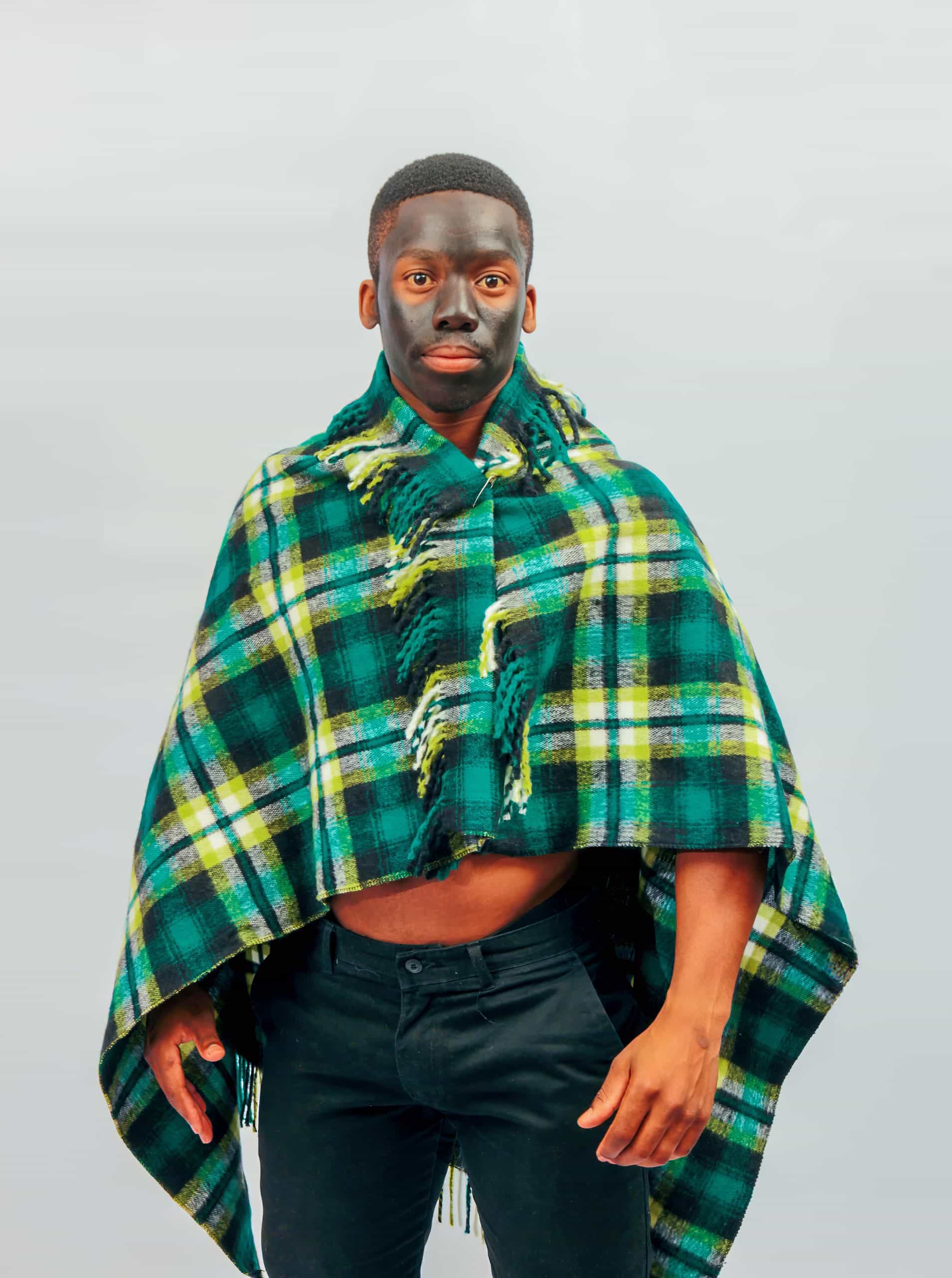 A man stands against a light background wearing a green, yellow, and blue checked poncho. His face has a pattern with areas of darker make-up contrasting his natural skin. He has short hair and is dressed in black trousers, exposing part of his midriff.