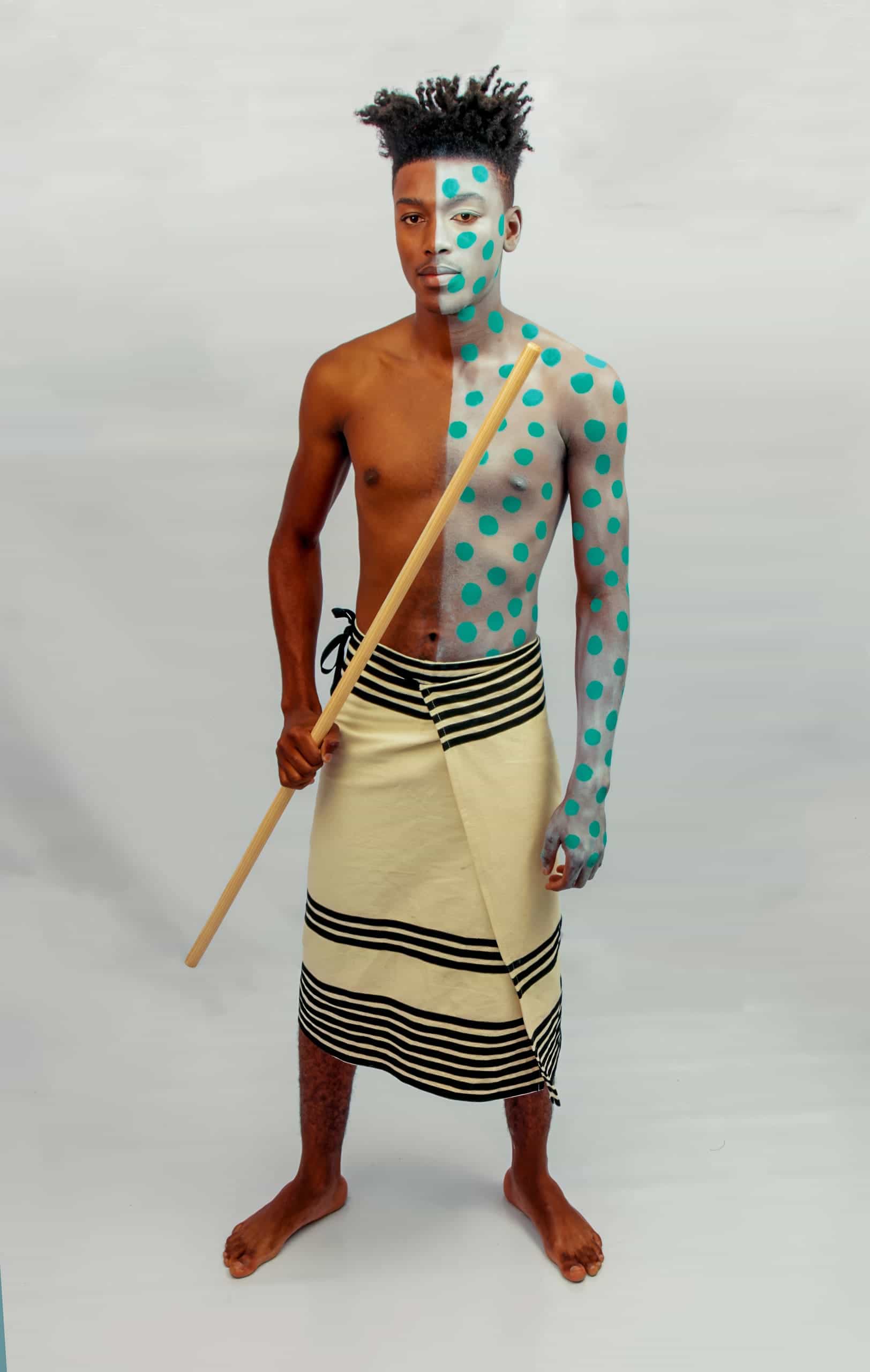 A barefoot man stands against a plain white background, wearing a cream-coloured wrap with black stripes. His upper body and face are painted half blue and half white, with blue polka dots on the white side. He holds a wooden staff vertically in one hand.