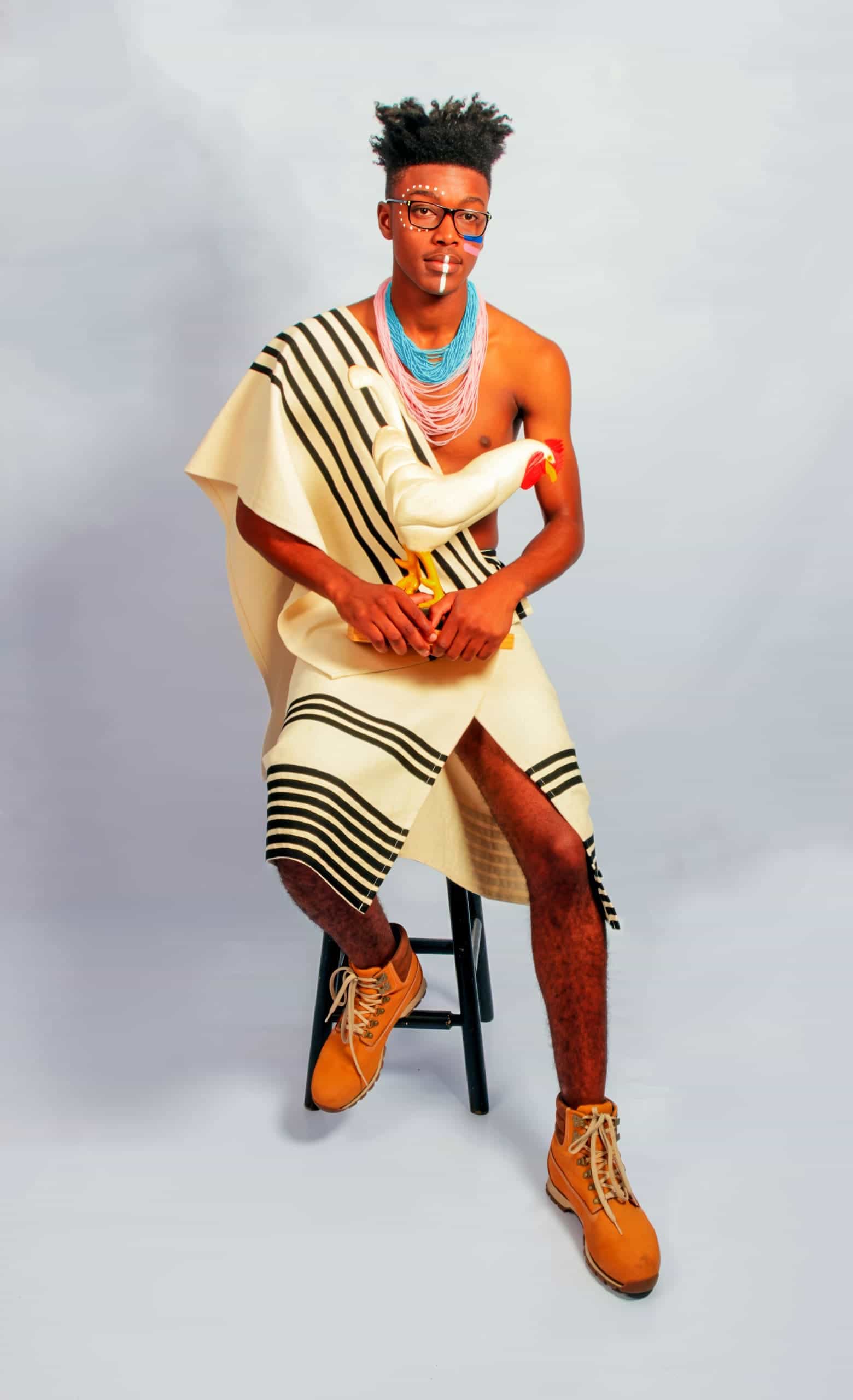 A man with glasses and beaded necklaces sits on a stool against a light background, draped in a cream and black striped cloth. He holds a white rubber chicken and wears tan boots. His expression is calm and he gazes directly at the camera.