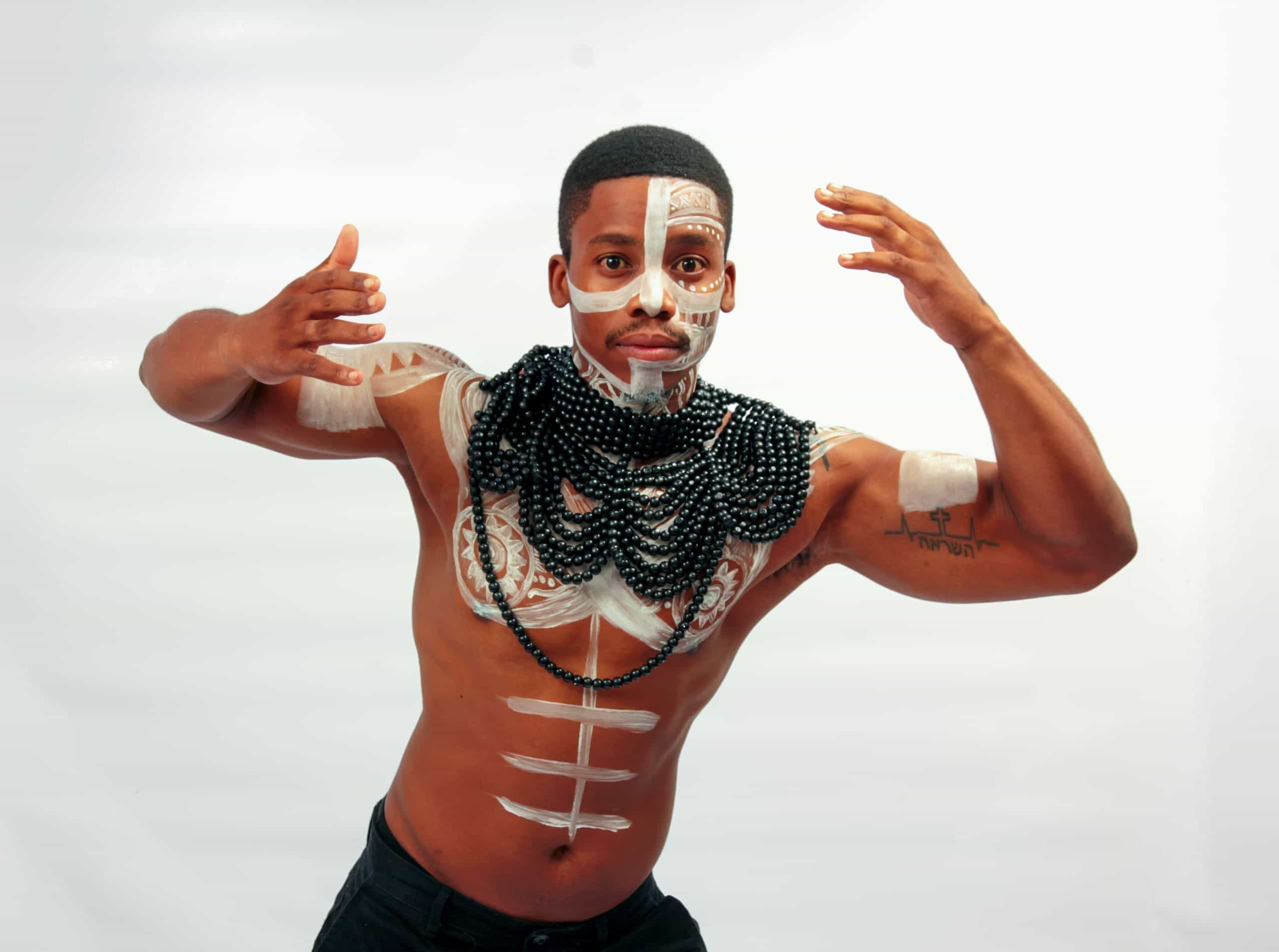A bare-chested man poses with arms raised, wearing black beaded necklaces and white tribal body paint on his face, chest, and arms. He stands against a plain white background, looking directly at the camera with a serious expression.