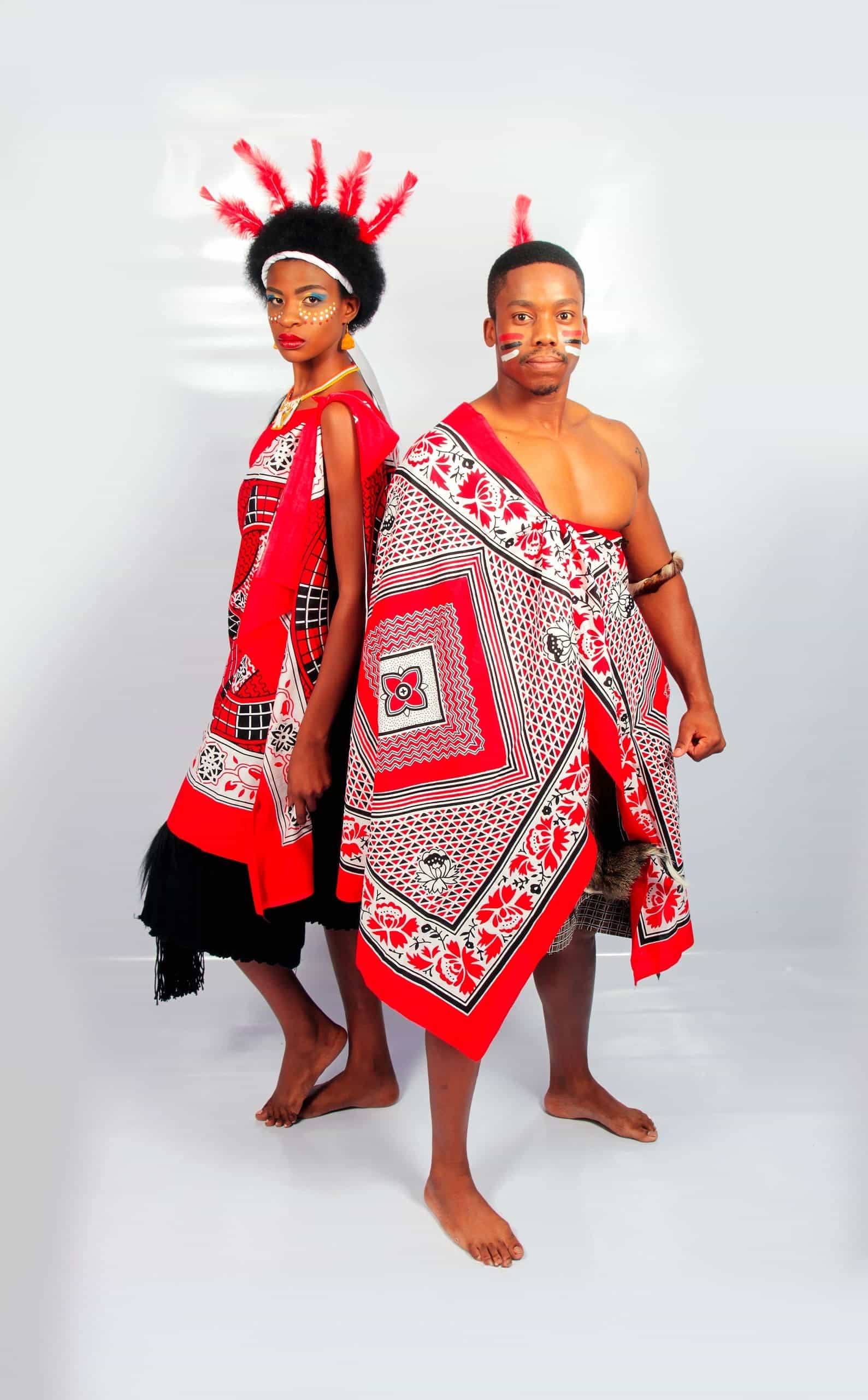 Two people wear vibrant red, black, and white traditional Swazi attire. The woman has face paint, a headdress with red feathers, and a patterned wrap. The man is bare-chested, with facial markings and a matching patterned cloth draped over his shoulder.