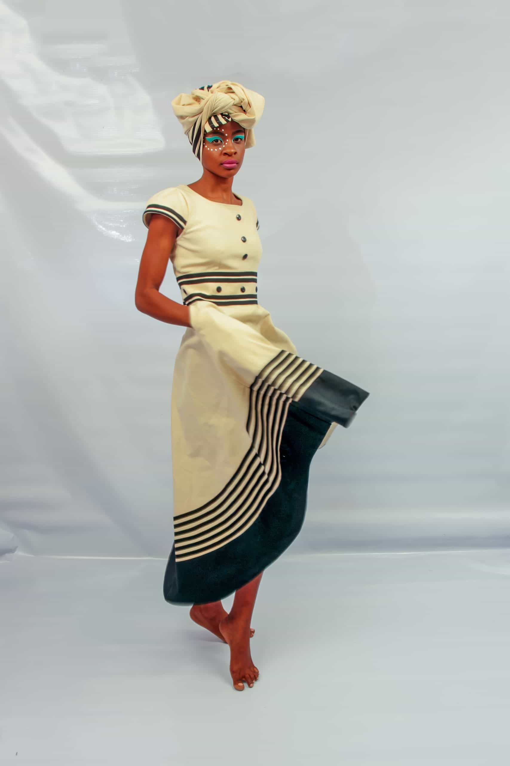 A person stands barefoot against a plain light backdrop, wearing a cream and black traditional dress with striped details. They have a matching headwrap and colourful face paint, looking directly at the camera whilst holding the skirt with one hand.
