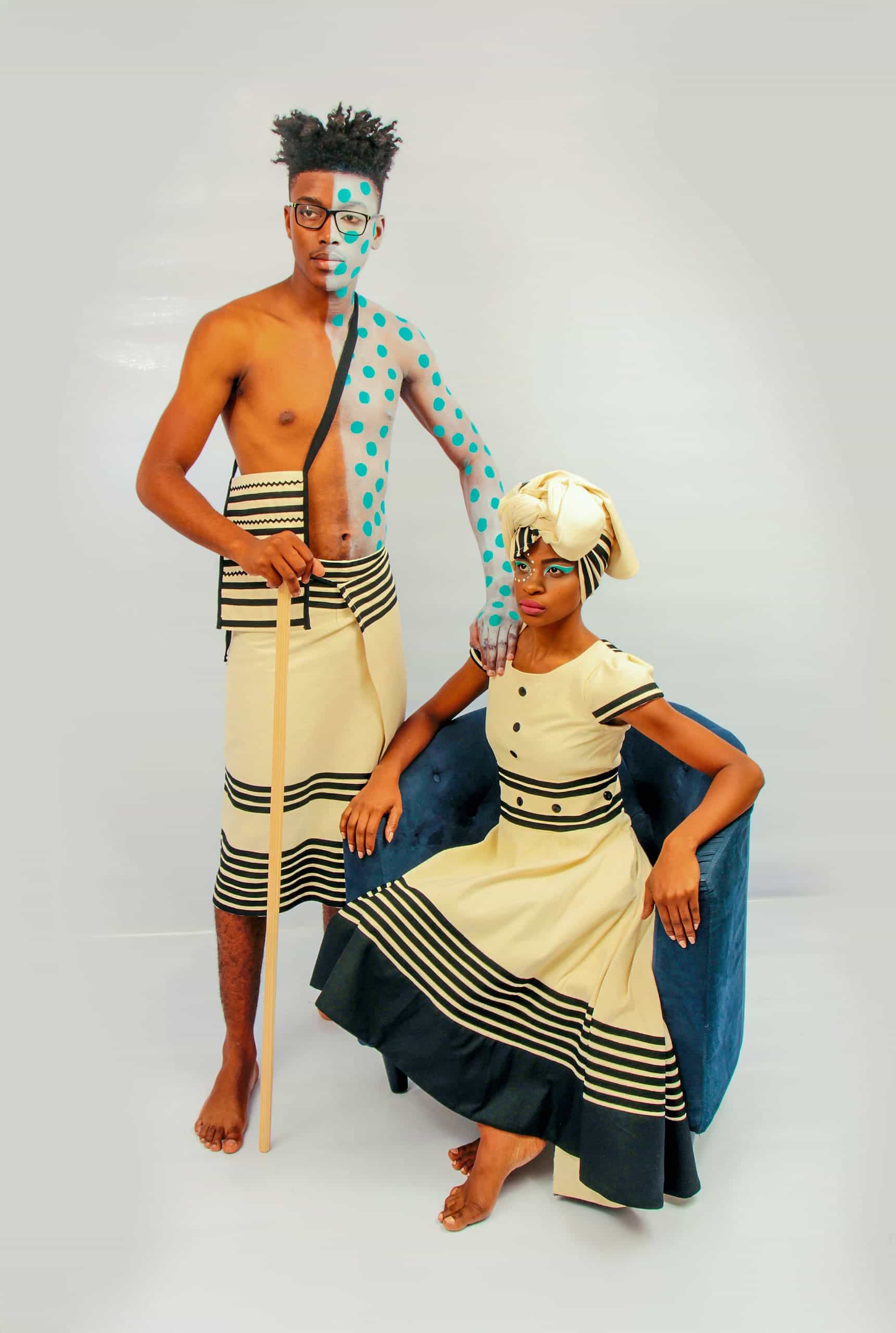 A barefoot man and woman pose in matching beige and black traditional attire. The man stands holding a wooden staff, his face and torso painted with turquoise dots and lines. The woman sits on a blue chair with a matching headwrap and face paint, both looking serious.