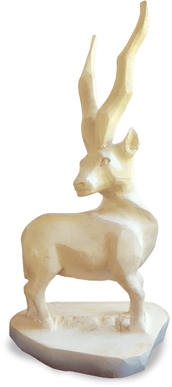 A light-coloured, roughly-carved wooden sculpture of an antelope stands on a flat base. The antelope has long, spiralled horns and is depicted in a static pose, facing left. The background is plain and has shades of beige and black.
