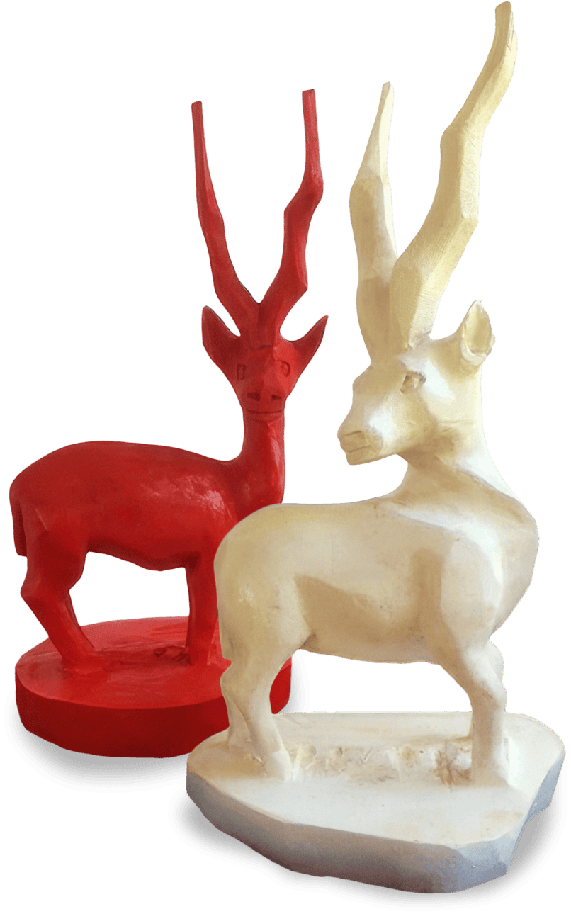 Two stylised antelope sculptures stand side by side. The figure in front is off-white with long, curved horns and angular features. The figure behind is bright red, slightly smaller, with similar elongated horns and a rounded base.