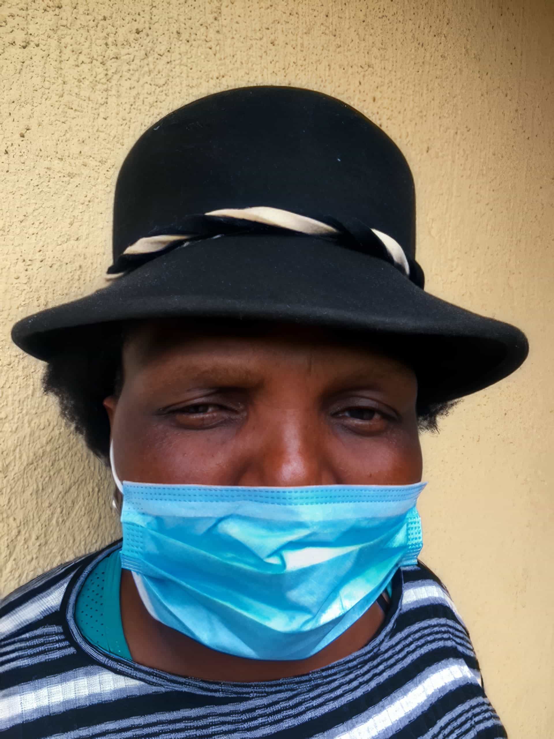 A person wearing a black hat with a white-and-black band and a striped shirt is facing the camera. They have a blue surgical mask covering their nose and mouth. The background is a beige textured wall.