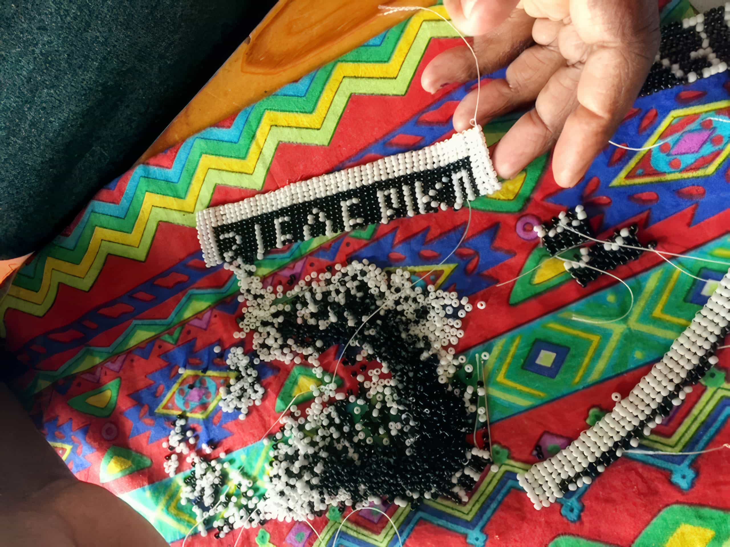 A hand holds a beaded craft spelling “AFRICA” in black and white beads on a colourful fabric with geometric patterns. Loose black and white beads and partially finished beadwork are scattered on the bright, patterned surface.
