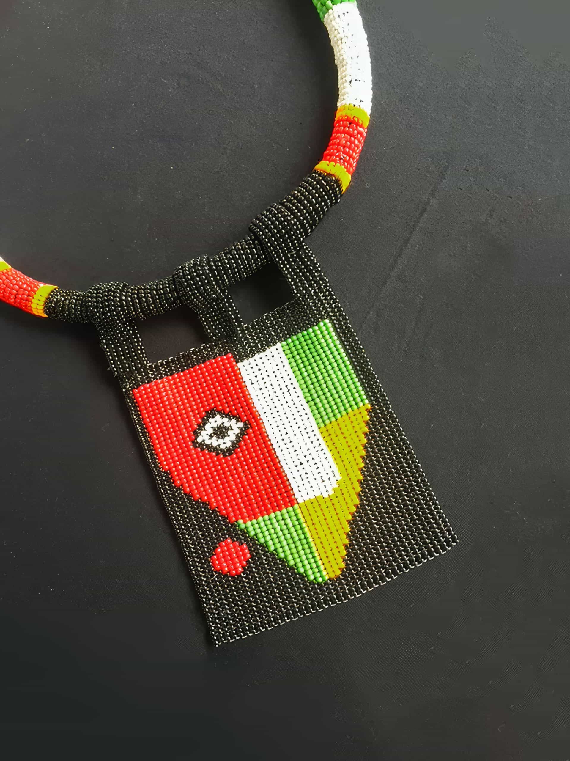 A beaded necklace features a rectangular pendant with a geometric design in red, white, green, yellow, and black beads. The pattern resembles the Togolese flag, with a red section containing a white star and various coloured sections. The necklace is on a dark surface.