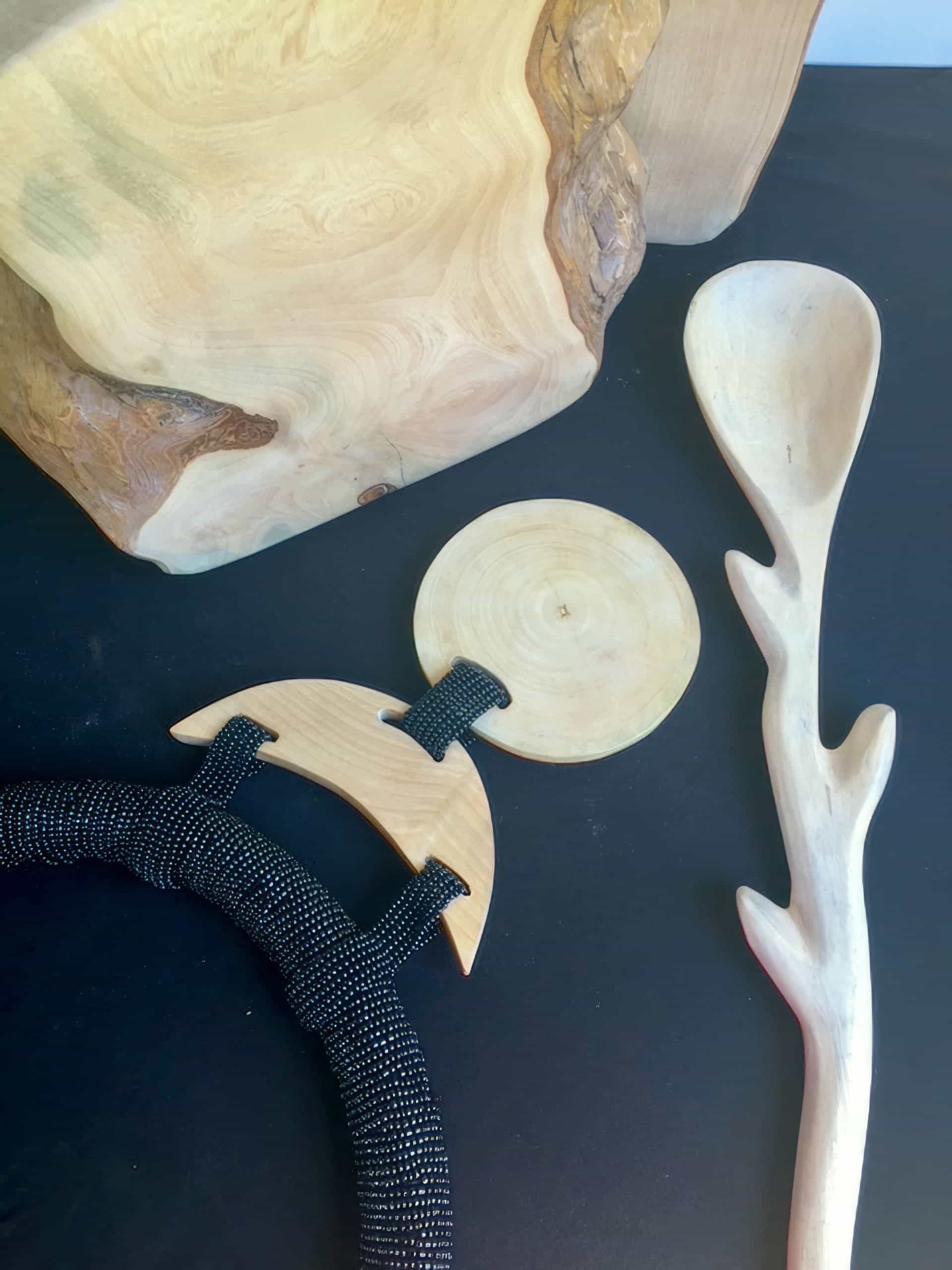 A photo shows a set of hand-carved wooden objects on a black surface: a large, blocky wood piece, a smooth circular disc, a crescent-shaped item with black rope, and a spoon with a handle shaped like a tree branch.