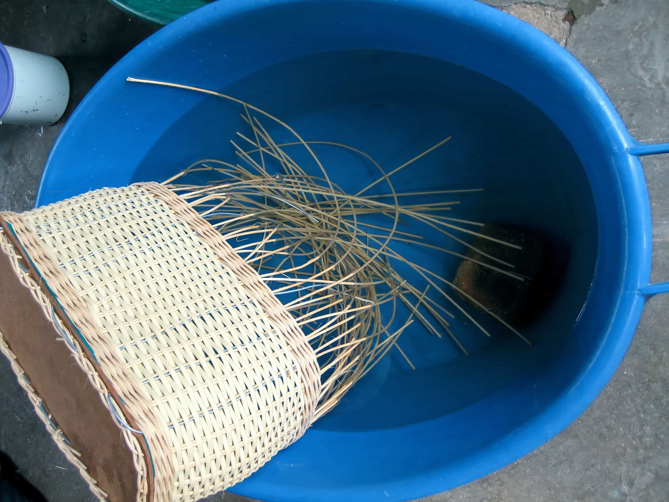 A partially woven wicker basket is soaking in a large blue plastic tub filled with water. Loose wicker strands extend from the unfinished basket, which rests inside the tub on a concrete floor. A sponge is also submerged in the tub.