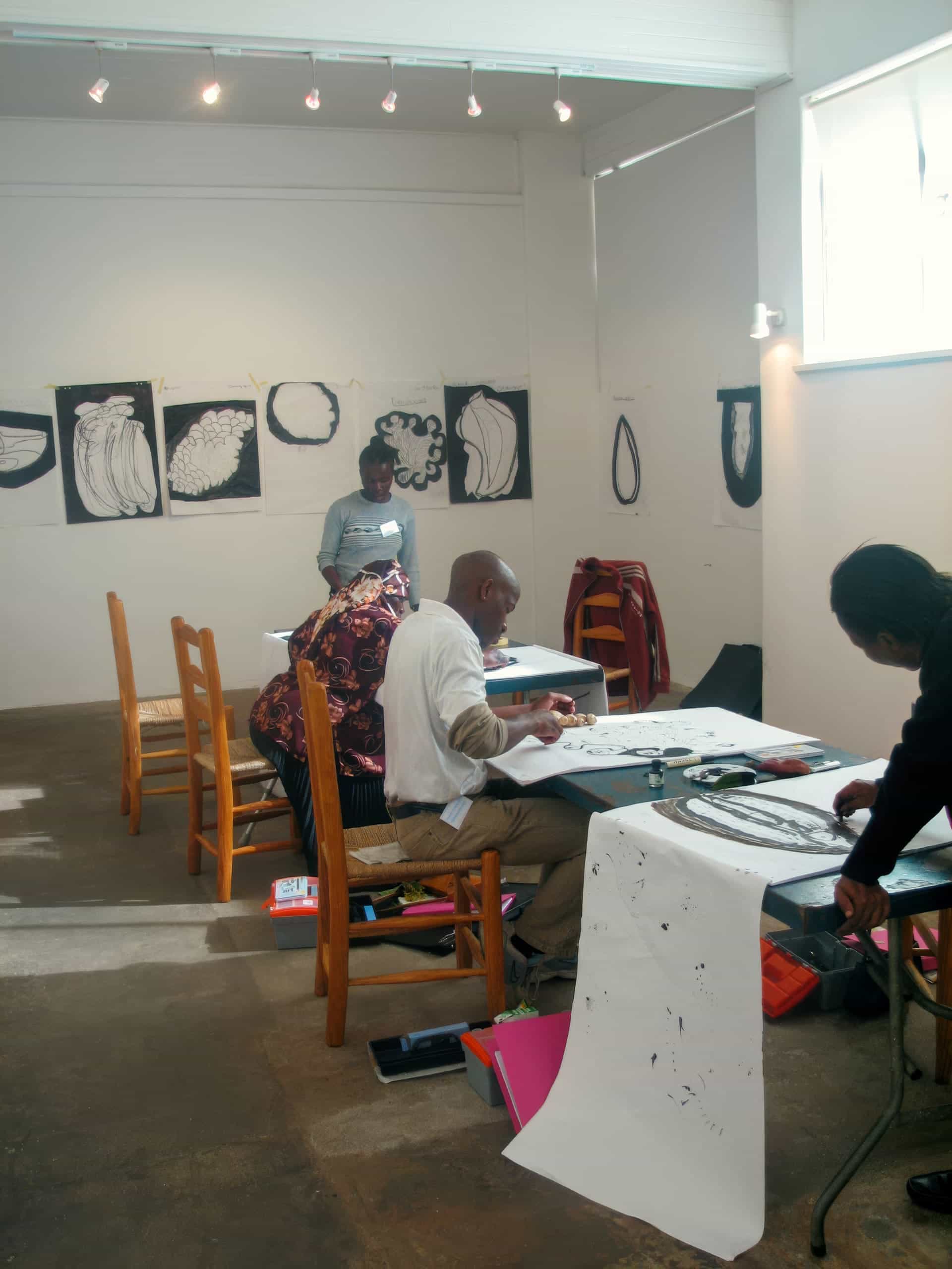 Three people sit at wooden tables in an art studio, sketching or painting on long sheets of paper. Black-and-white abstract art hangs on the walls. Another person stands in the background. The room is well-lit with natural and track lighting.