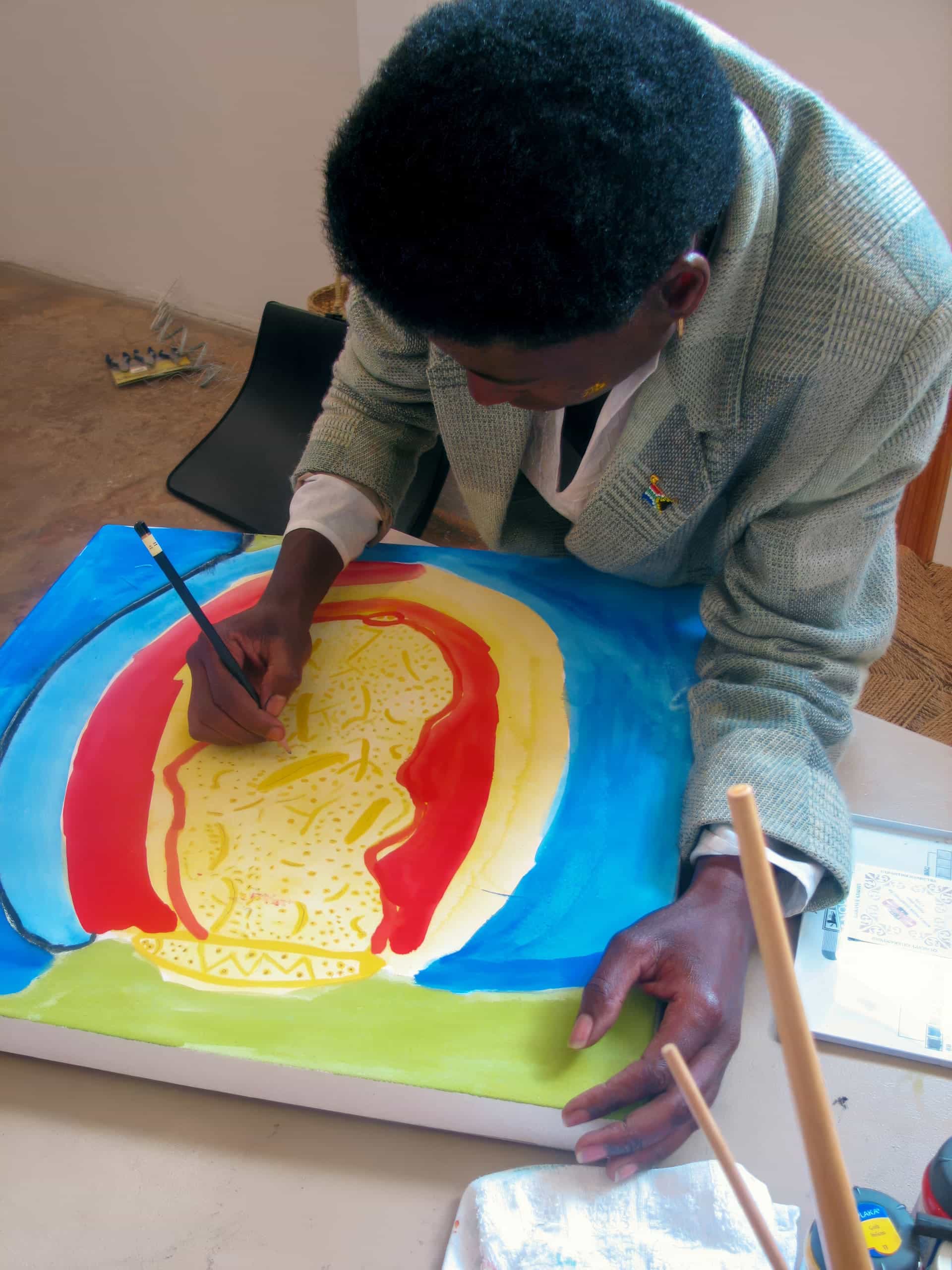 A person with short, curly hair and wearing a grey jacket is bending over a table, painting a colourful abstract image with yellow, red, and blue areas on a canvas. Art supplies and brushes are scattered on the tabletop in the foreground.